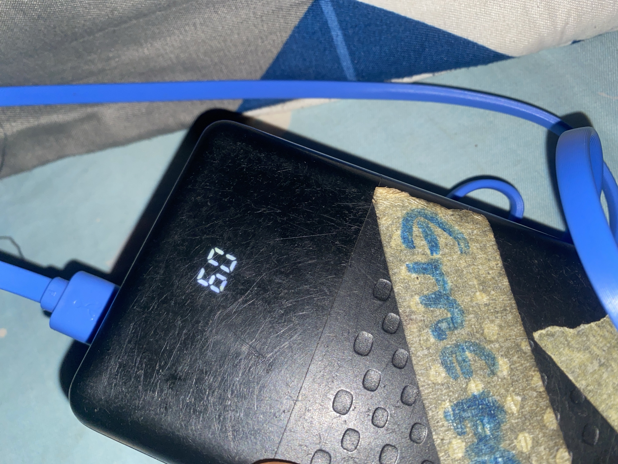 The image shows a close-up of a black power bank that is currently charging. A blue charging cable is plugged into the power bank. The power bank has a digital display showing "60", likely indicating the battery percentage. There are several small, circular raised dots on the surface of the power bank. A strip of tan-colored tape with blue writing is affixed to the power bank. The blue writing is partially obscured but appears to be a name or word, with letters like "Gree" visible. The power bank is placed on a surface that has a blue and white checkered pattern, possibly a cloth or bedding. The lighting suggests it is indoors, with no strong indications of the time of day or weather conditions. The overall impression is a functional object in a domestic setting.