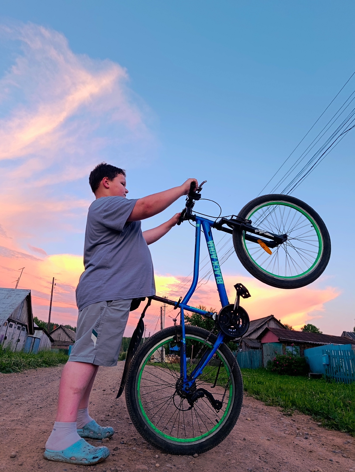 In Зипуново, Russia, a young boy stands on a dirt road, holding the handlebars of a blue bicycle upside down. The sky is a vibrant mix of blue and pink clouds, suggesting sunset. The boy wears a gray t-shirt and gray shorts, with light blue clogs and white socks. The bicycle has green rims on its wheels. In the background, there are wooden houses, a blue fence, and green grass. Electrical wires are visible against the sky. The scene appears to be in a rural setting.