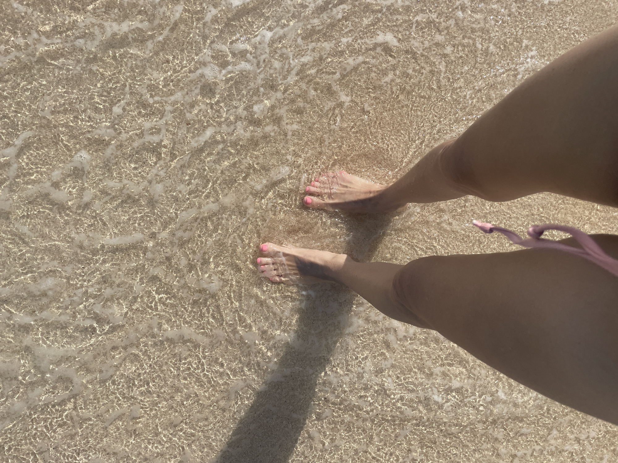 A first-person perspective shot of a person's legs and feet standing in shallow, clear ocean water on a sandy seabed. The water has ripples and reflections from the sunlight, creating a textured pattern on the sand. The person's feet are positioned in the center of the frame, with toes pointing slightly upwards. Their toenails are painted a bright pink color. A shadow of the person's left leg is cast diagonally across the sand to the left of the frame. Part of a pink bikini string is visible at the bottom edge of the image. The location is Kuta Selatan, Indonesia, implied by the tropical beach setting and clear, warm water.