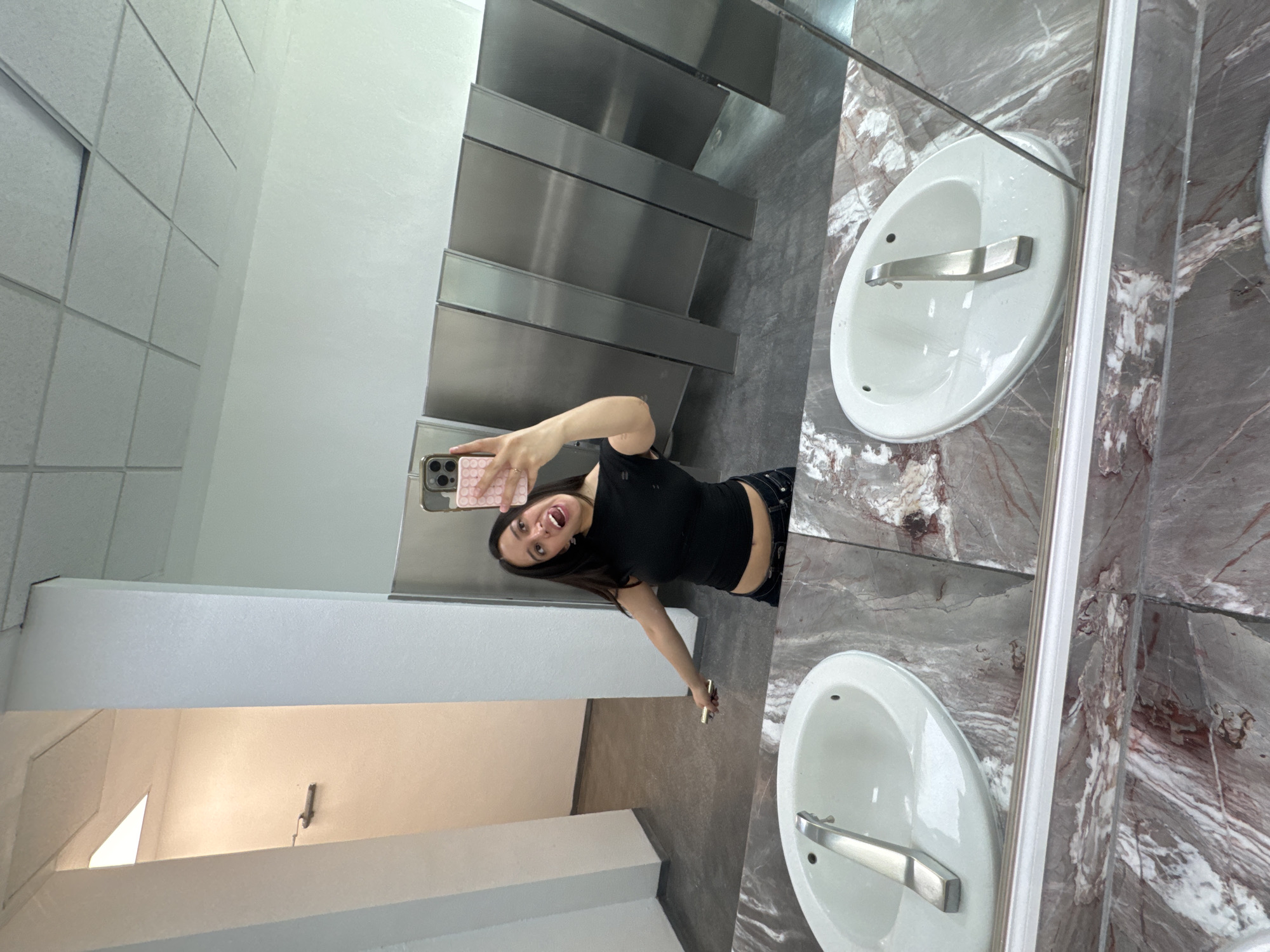 A young woman is reflected in a mirror in a public restroom. She is holding a cell phone in her left hand, presumably taking a selfie. She is wearing a black tank top and dark pants. The restroom has multiple stalls with stainless steel doors. The walls are white. There are two white sinks with silver faucets and marble countertops visible in the mirror. The location is Puebla, Mexico.