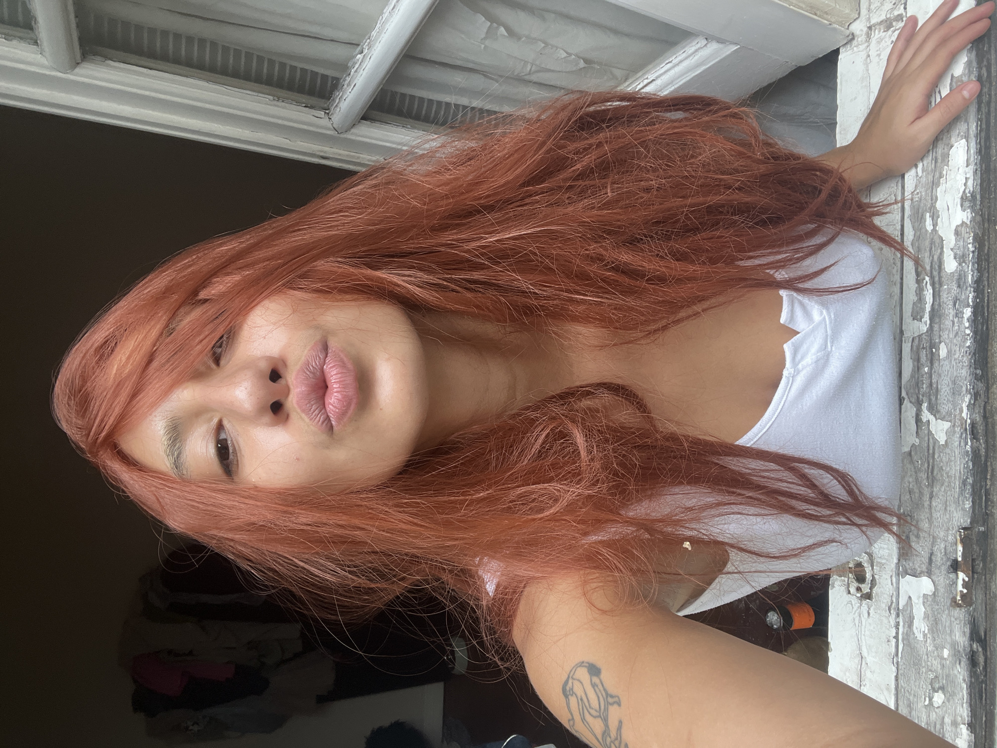 The image features a young person with long, flowing reddish-brown hair, shown from the chest up. Their face is turned towards the camera, adorned with a playful "duck face" or pouting expression, with lips slightly parted. Their right eye is partially visible, while the left is mostly obscured by hair. A prominent tattoo depicting an abstract or stylized creature, possibly marine life, is visible on their right forearm. The person is wearing a simple white tank top.
The subject is positioned within what appears to be an old window or an opening in a wall, characterized by a thick wooden frame with heavily peeling white paint, indicating significant wear and age. Their right hand rests casually on this distressed frame. The background visible through the opening is dark and indistinct, offering little detail beyond shadowed shapes. The lighting is soft and even, highlighting the subject's features and hair.
The image captures a casual, posed moment, likely a selfie. The setting, given the context, is Buenos Aires, Argentina, and the weathered appearance of the window frame could be indicative of older architecture common in the city. The overall mood is relaxed and confident. No discernible text is visible within the frame.