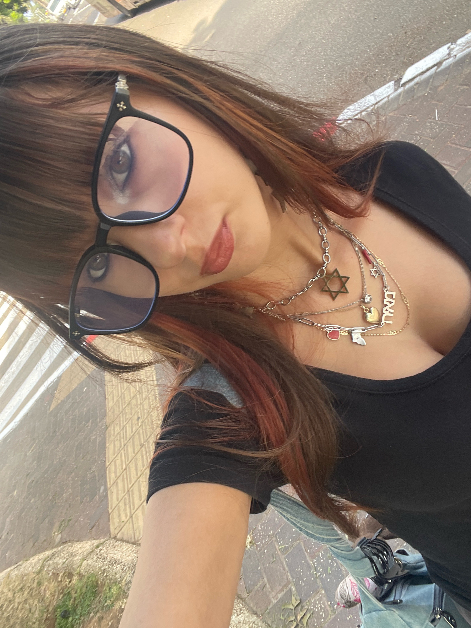 In Raanana, Israel, a woman with long, dark brown hair with red highlights is captured in a selfie taken outdoors. She is wearing glasses with black frames, a black t-shirt, and multiple layered necklaces. One necklace features a Star of David pendant, and another has a pendant with Hebrew lettering that appears to spell "CHAI." The background shows a paved sidewalk with a white line and a street. A portion of a vehicle is visible in the upper left corner.