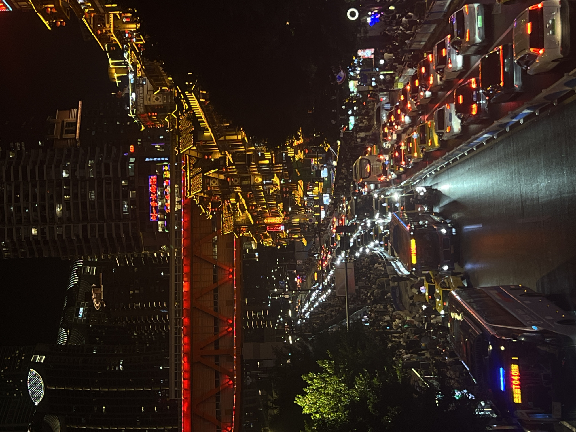 The media file depicts a bustling city scene at night, likely in Yuzhong District, China, characterized by vibrant lights and heavy traffic. The perspective is from an elevated viewpoint, looking down upon a densely populated urban environment.
The primary subjects are a multitude of cars and buses lining a road, many with their headlights and taillights illuminated, indicating a significant traffic jam. The vehicles are closely packed, suggesting congestion. Numerous people are visible, either walking along the streets, gathered on sidewalks, or possibly in viewing areas, their presence contributing to the lively atmosphere.
The scene is set against a backdrop of towering buildings, many adorned with bright, decorative lights, showcasing a mix of modern architecture and traditional-inspired designs, particularly on what appears to be a pedestrian street or market area. The overall ambiance is one of activity and festivity, with the abundance of lights suggesting a special occasion or simply the energetic nightlife of a major city.
Notable details include the time of day, which is clearly night, as indicated by the darkness and the prevalence of artificial lighting. There are no obvious cues about the weather, but the scene suggests a clear night. The abundance of lights, the crowds, and the traffic point towards a lively urban environment.
Visible text includes Chinese characters on signs and on the vehicles. For instance, some vehicles have illuminated text in Chinese, such as "0401" on one of the buses. The architectural elements also display illuminated signage, though specific characters are not clearly legible in all instances. The overall impression is a dynamic and vibrant urban landscape at night, filled with people and vehicles.