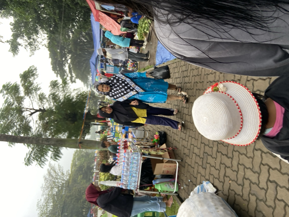 The image shows a street market in Bandung, Indonesia, located at coordinates -6.896 latitude and 107.619 longitude. The market is held on a paved street with brick-like blocks, lined with trees and vendors. A young girl wearing a white sun hat is in the foreground. Two women are walking towards the camera, one wearing a blue patterned headscarf, a blue patterned long dress, and a black plastic bag, and the other woman wearing a black headscarf, a yellow long-sleeved shirt, and black pants. There are other people walking around the market and behind the two women in the foreground. Vendors are seen setting up their stalls along the side of the street. The scene appears to be a typical street market in a bustling city. The weather is cloudy, and it appears to have been raining recently.