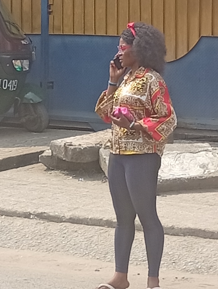 An adult female is depicted standing outdoors in Umuode, Nigeria, engaged in a phone call. She possesses dark, curly hair adorned with a red fabric headband tied into a bow. Her face is partially obscured by pink-tinted sunglasses, and she holds a dark-colored smartphone to her right ear with her right hand. On her left wrist, she wears a light blue bracelet and another light-colored bracelet on her right wrist. Her attire consists of a long-sleeved, patterned button-up shirt featuring gold, white, and red baroque-style motifs, paired with dark grey form-fitting leggings and white flat sandals. In her left hand, she holds a small, bright pink, soft-textured pouch or bag.
The subject stands on an uneven, light-colored concrete or paved surface. To her immediate right, several irregularly shaped concrete blocks or pieces of rubble are visible on the ground. In the background, partially visible on the left, is the front left section of a maroon-colored auto-rickshaw (tuk-tuk) displaying a white plate with "0419". Behind the vehicle and the woman is a blue metal gate or wall, with a section of a light brown or yellowish vertical slat fence visible above it. The scene is illuminated by bright daylight.