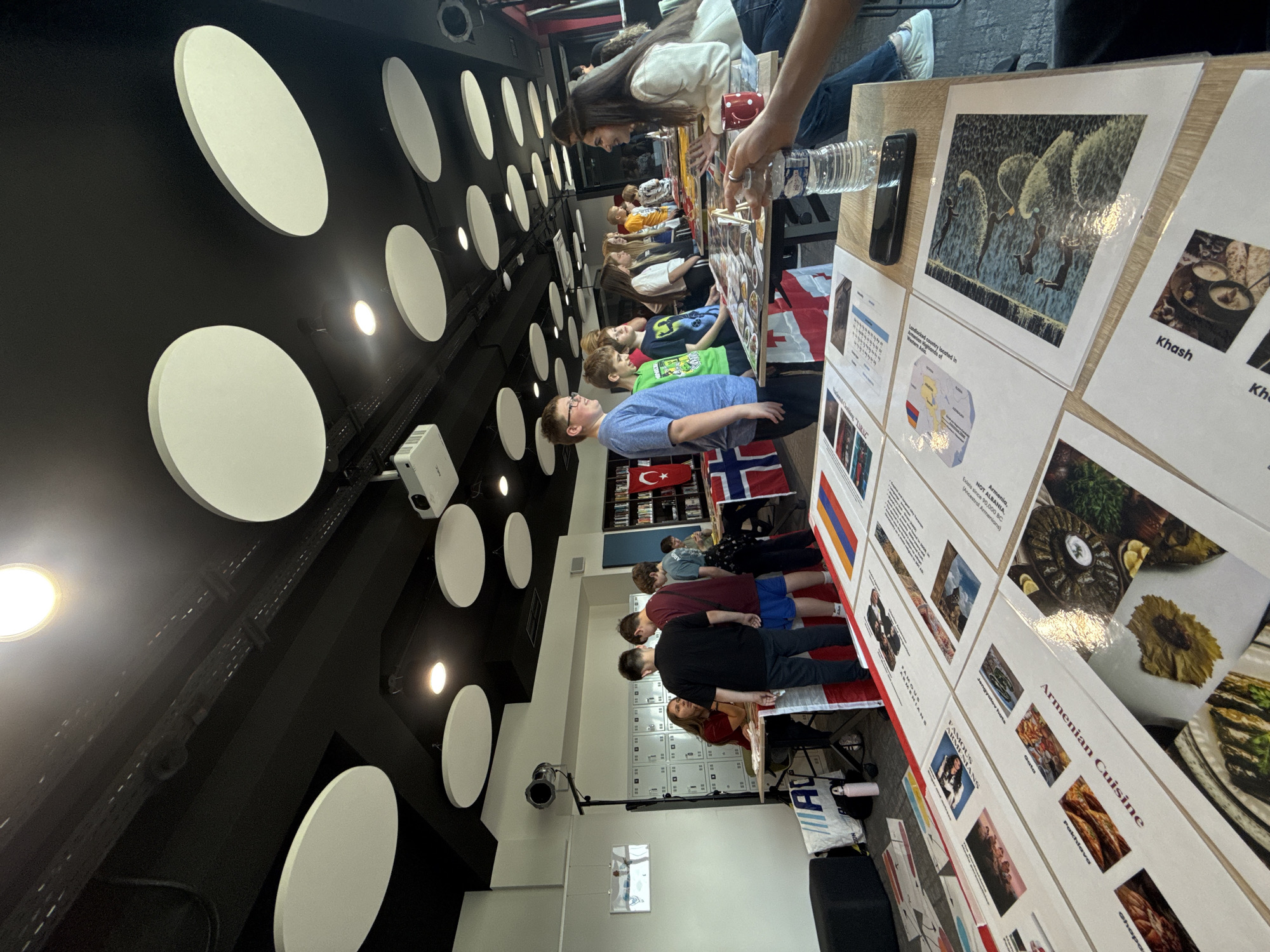 In the image, a cultural fair is taking place. Several people, including young adults and a woman, are interacting around tables displaying information and images related to various countries, with flags visible, including those of Armenia, Norway, and Turkey. The environment appears to be indoors, possibly a school or community center, with a distinctive ceiling featuring large, circular acoustic panels and spotlights. The tables are covered with printed materials, some showcasing "Armenian Cuisine" and other cultural exhibits. The overall atmosphere suggests an educational event promoting international understanding and cultural exchange. The scene is set in the city of Opole, Poland.