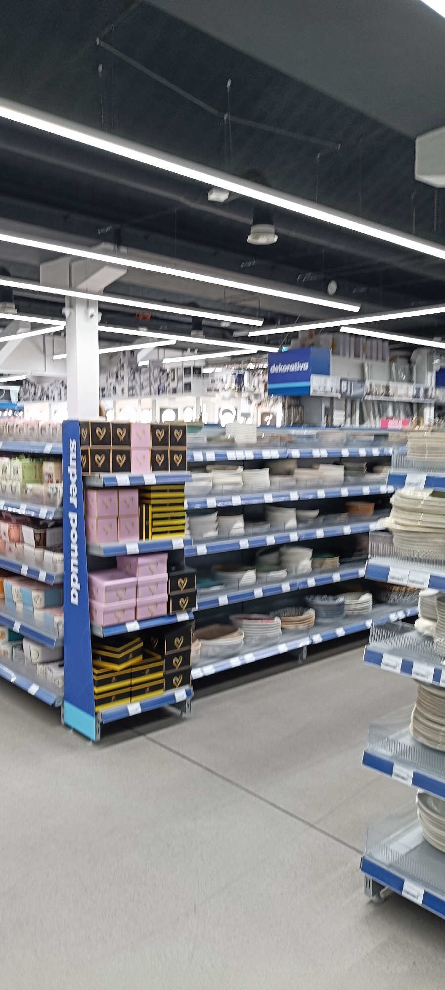 The image depicts the interior of a well-lit retail store, likely a home goods or kitchenware store, in Novi Sad, Serbia. The scene is dominated by rows of shelves filled with various products. On the left, a blue display pillar with the text "SUPER POMUDA" is visible, featuring stacked boxes decorated with heart patterns. Adjacent shelves display more boxed items, including pink and gold boxes, and yellow striped boxes. Further to the right, numerous shelves are stacked with white plates and bowls, some with intricate patterns, and decorative items. In the background, a sign with the word "dekorativa" in blue lettering can be seen, suggesting a decorative items section. The lighting appears to be from overhead fluorescent fixtures. The overall impression is one of a retail environment stocked with merchandise, ready for customers. There are no people visible in the image. The floor is a light gray, and the overall atmosphere suggests a typical weekday shopping experience.