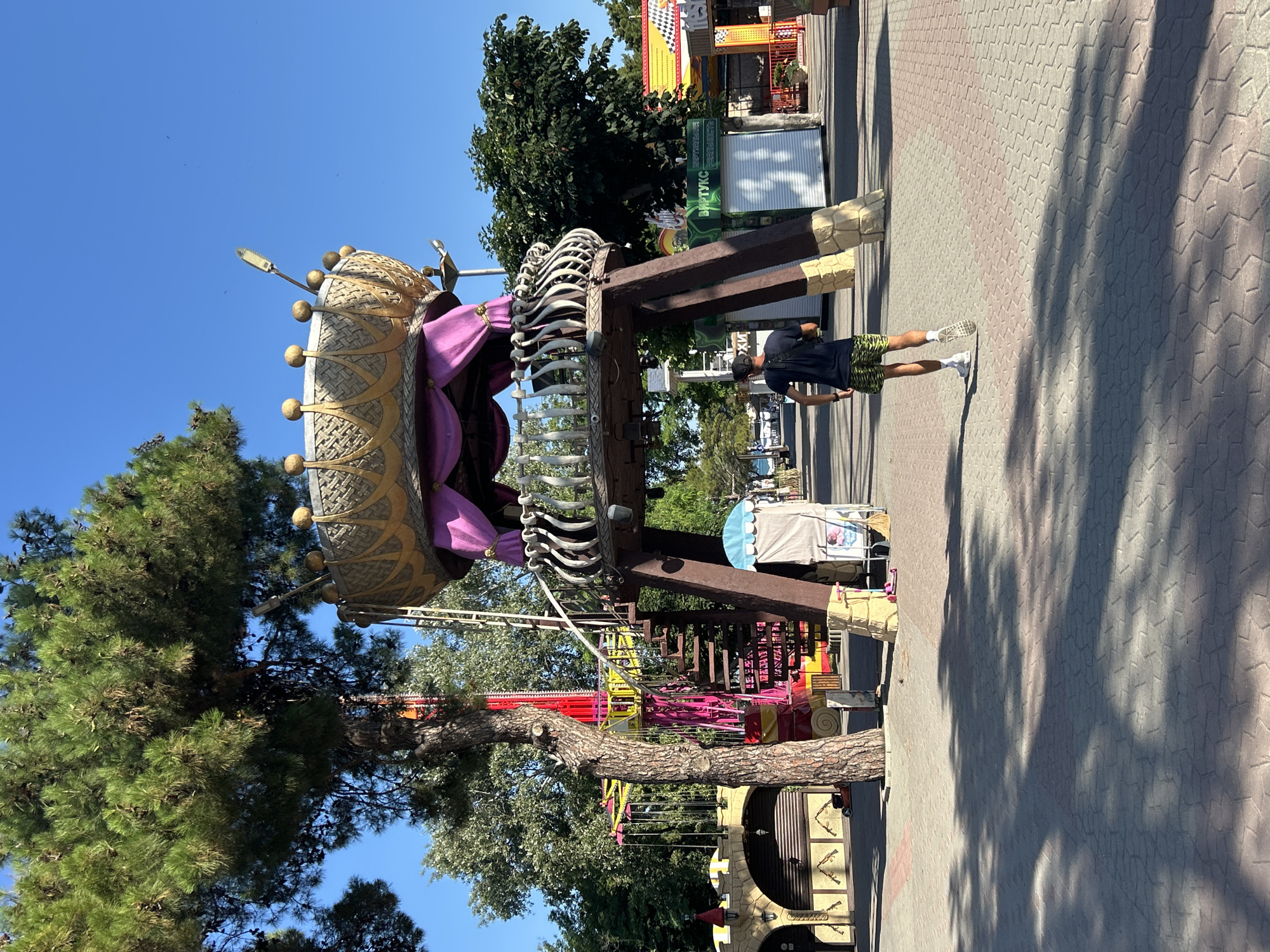 The image captures a sunny day in an amusement park in Gelendzhik, Russia. In the foreground, a person is walking away from the camera on a patterned brick pathway. The person is wearing a dark t-shirt, camouflage shorts, white socks, and white sneakers. Behind the person, a large, ornate structure resembling a crown sits atop thick wooden legs. This structure is decorated with purple fabric and golden accents. To the left of this structure, a tall, red and yellow swing ride is visible, with a spiral staircase leading up to the crown-like attraction. Further back, various kiosks and small buildings line the pathway, suggesting a commercial area within the park. Trees and foliage are interspersed throughout the scene, and a clear blue sky dominates the upper portion of the image. The overall impression is that of a vibrant and active amusement park.