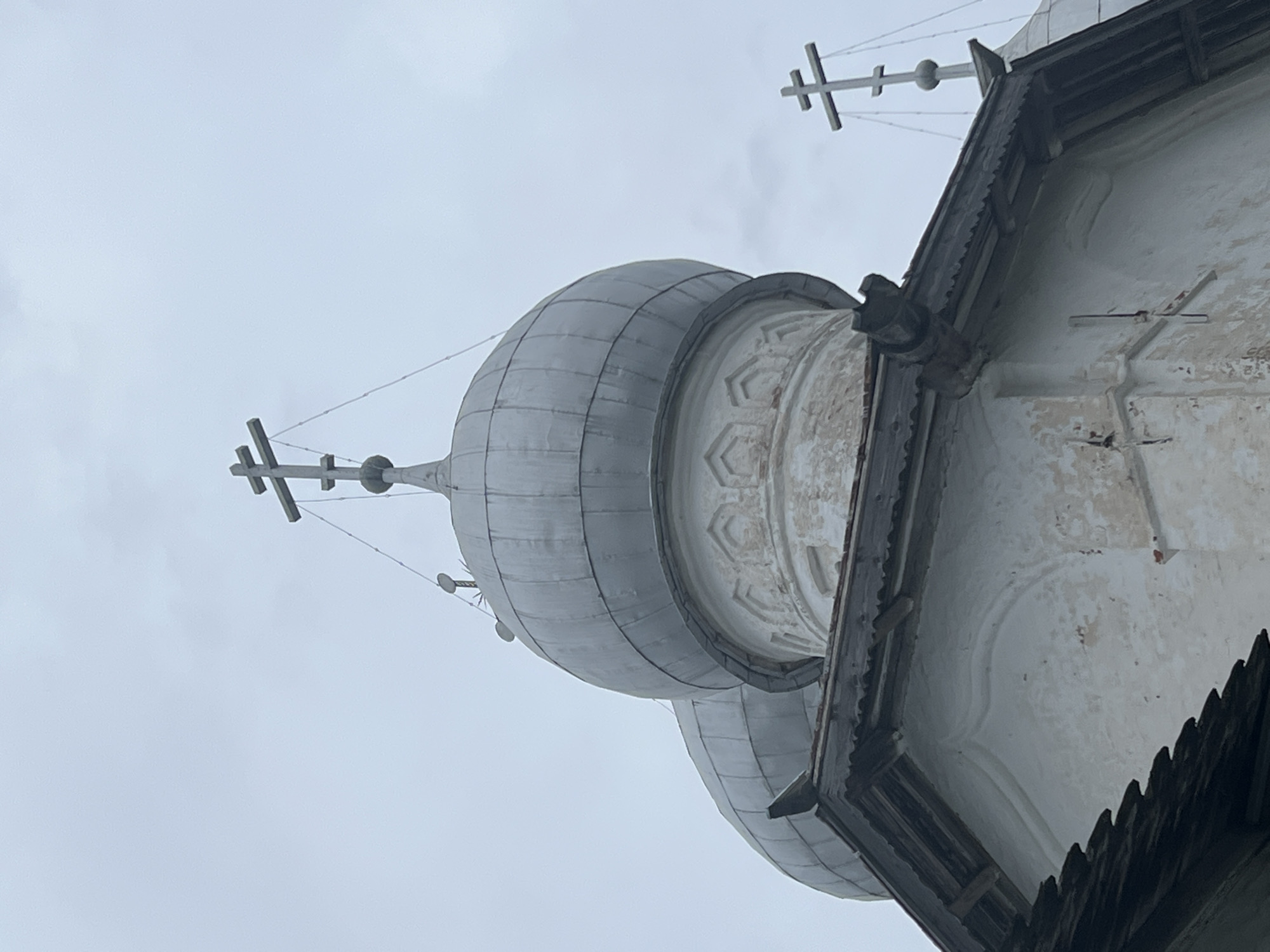 The image shows the upper portions of several domes and roofs of a historic building, likely a church, against an overcast sky. The domes are metallic and rounded, topped with multi-barred Orthodox crosses. The building itself appears to be made of white plaster with visible signs of age and wear, including cracks and discolored patches. There are wooden architectural elements, such as a decorative eave line with carved trim. The scene is devoid of people or any discernible activity. This architecture is consistent with the historical religious buildings found in Veliky Novgorod, Russia.