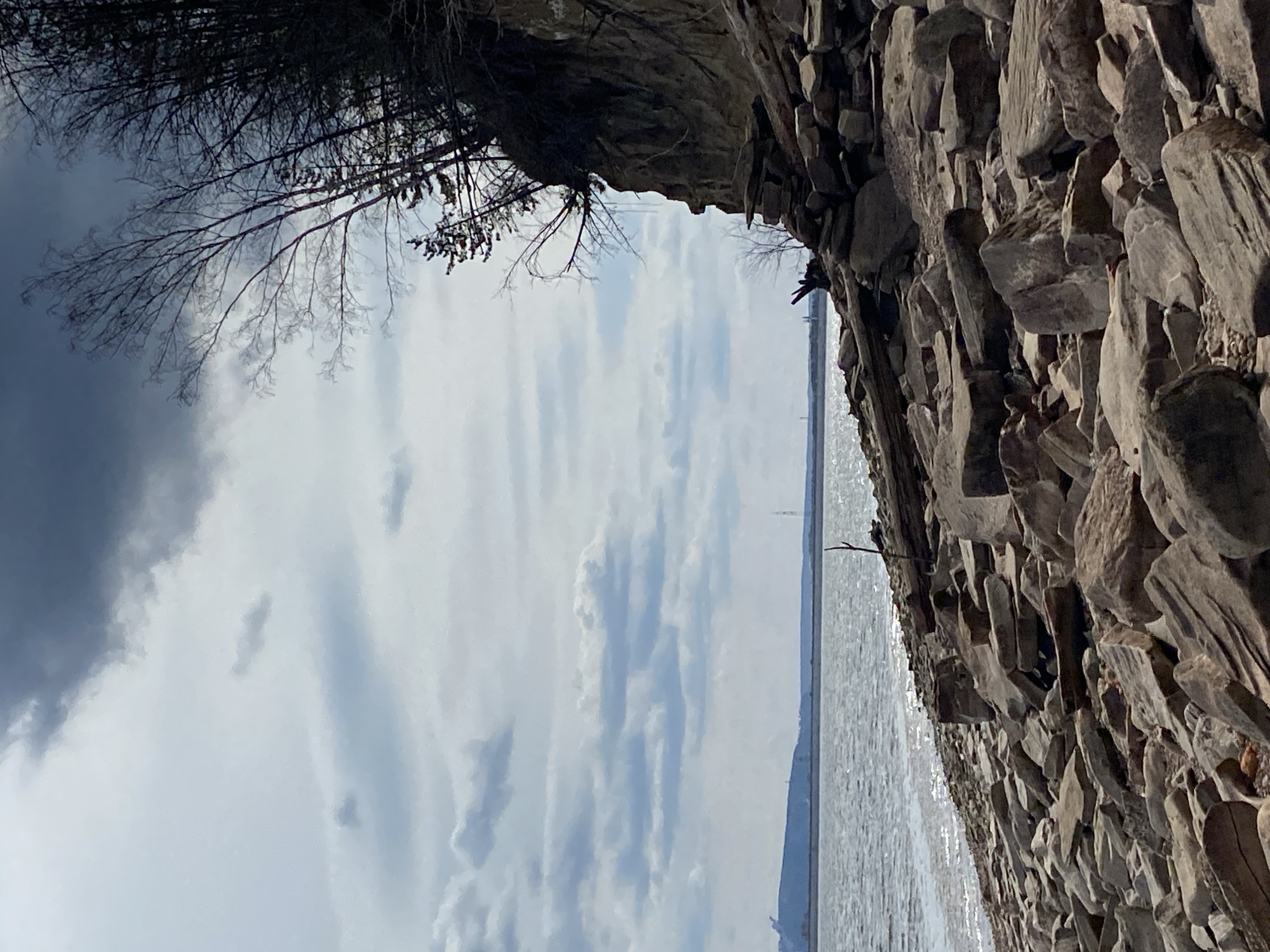 A landscape photograph taken on a cloudy day captures a waterscape scene from a low-angle perspective, seemingly from within a grotto or cave-like structure. The foreground is dominated by a rough, rocky wall composed of various sized stones, stacked and cemented together. These stones appear light gray to brownish in color, indicating a natural or possibly constructed wall facing a body of water.
Above the rock wall on the left, bare tree branches reach into the sky, silhouetted against the cloudy expanse. The sky is a mix of dark and light gray clouds, hinting at an overcast or potentially stormy weather condition.
Beyond the opening, a body of water stretches into the distance. The water surface reflects the sky, creating a shimmering effect. In the distance, faint land features can be observed, suggesting a shoreline or distant hills.
Given the prompt's instruction, it is reasonable to infer that this scene is located in the city of Chaikovsky, Russia.