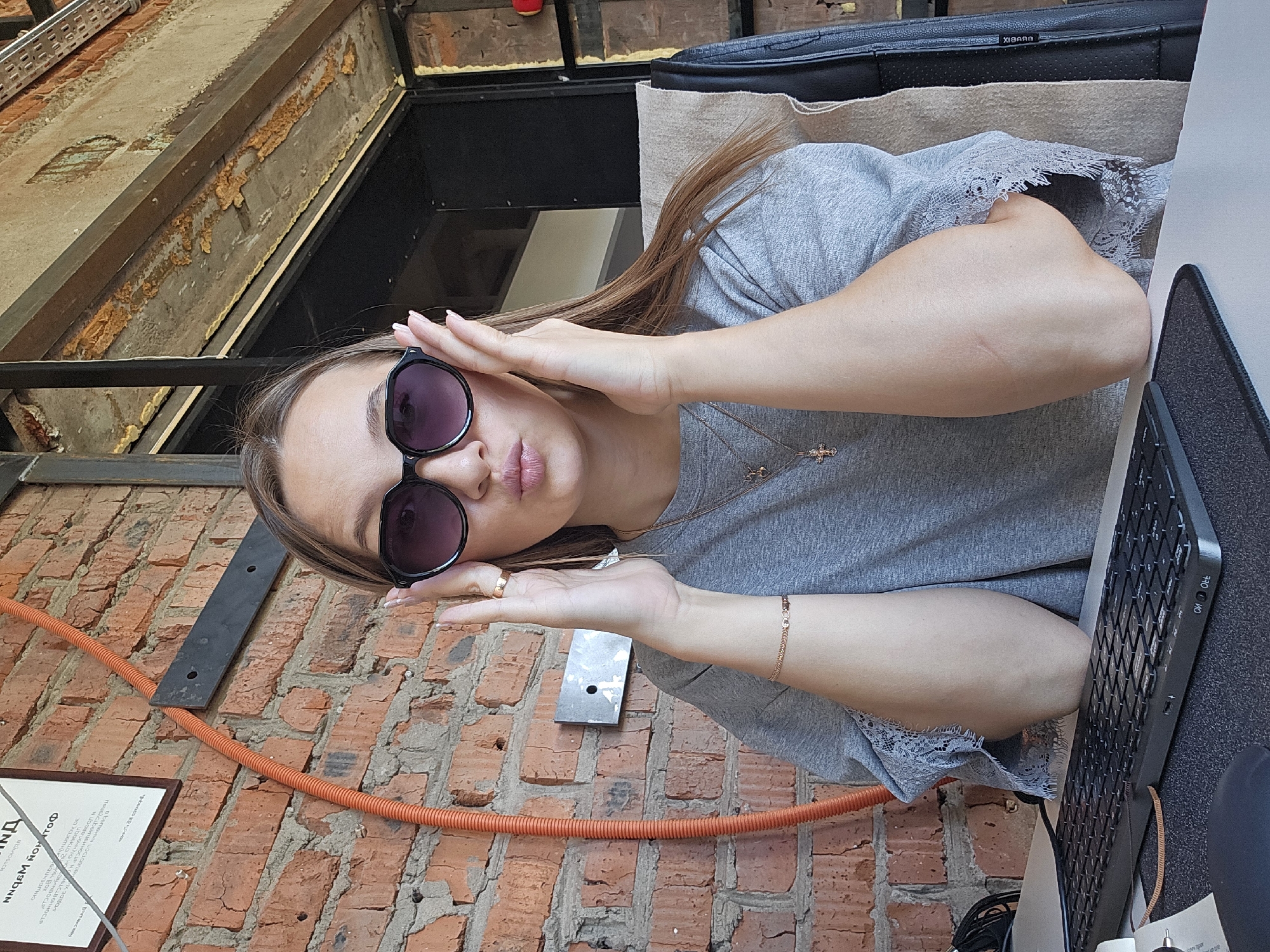 A young woman with blonde hair is sitting at a desk in an office space with exposed brick walls. She is wearing sunglasses and a grey t-shirt, and she has a pouty expression. Her hands are resting on her face, and she is wearing a bracelet and a necklace with a cross pendant. A keyboard is visible on the desk in front of her. The office appears to be industrial in style, with visible pipes and exposed ceiling beams. There is a framed document on the wall behind her, and an orange pipe runs vertically up the brick wall. The image appears to be a candid photograph taken in an office environment in Chaikovsky, Russia.