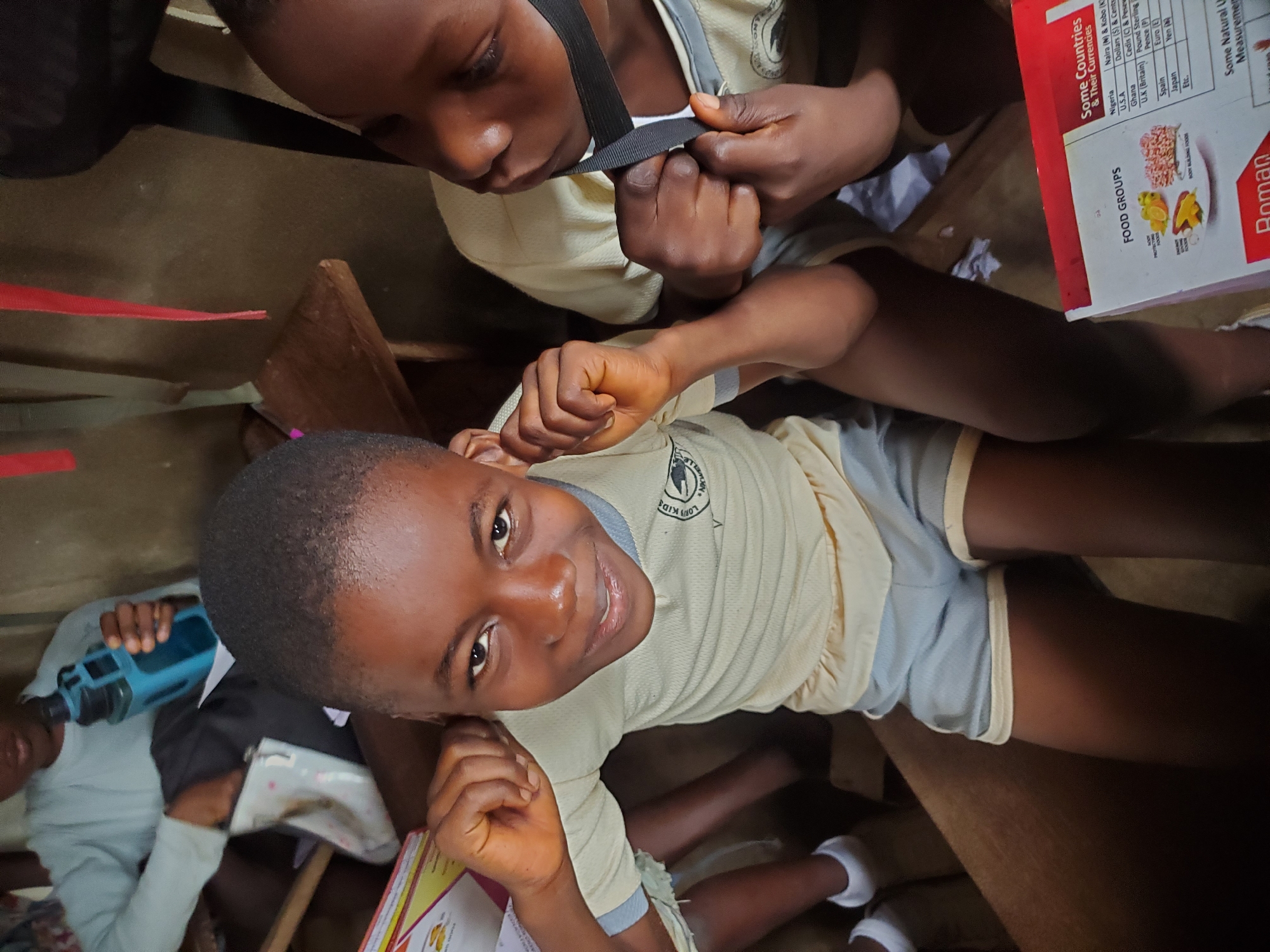 The image shows two children in what appears to be a classroom setting in Ukwu-Abwa, Nigeria. One child, positioned towards the bottom of the frame, is looking directly at the camera and smiling. This child is wearing a light yellow shirt with grey trim and grey shorts, indicating a school uniform. Part of a school logo is visible on the shirt. The child is seated at a wooden desk.
The second child is partially visible at the top of the frame, focused on adjusting a black strap, possibly part of a bag or uniform. This child is also wearing the same uniform shirt.
On the desk near the smiling child, there is an open textbook about nutrition and world currencies. The text and images indicate an educational context related to food groups and international finance.
In the lower-left corner of the image, part of a third person can be seen holding a blue object. The environment suggests a modestly furnished classroom interior.