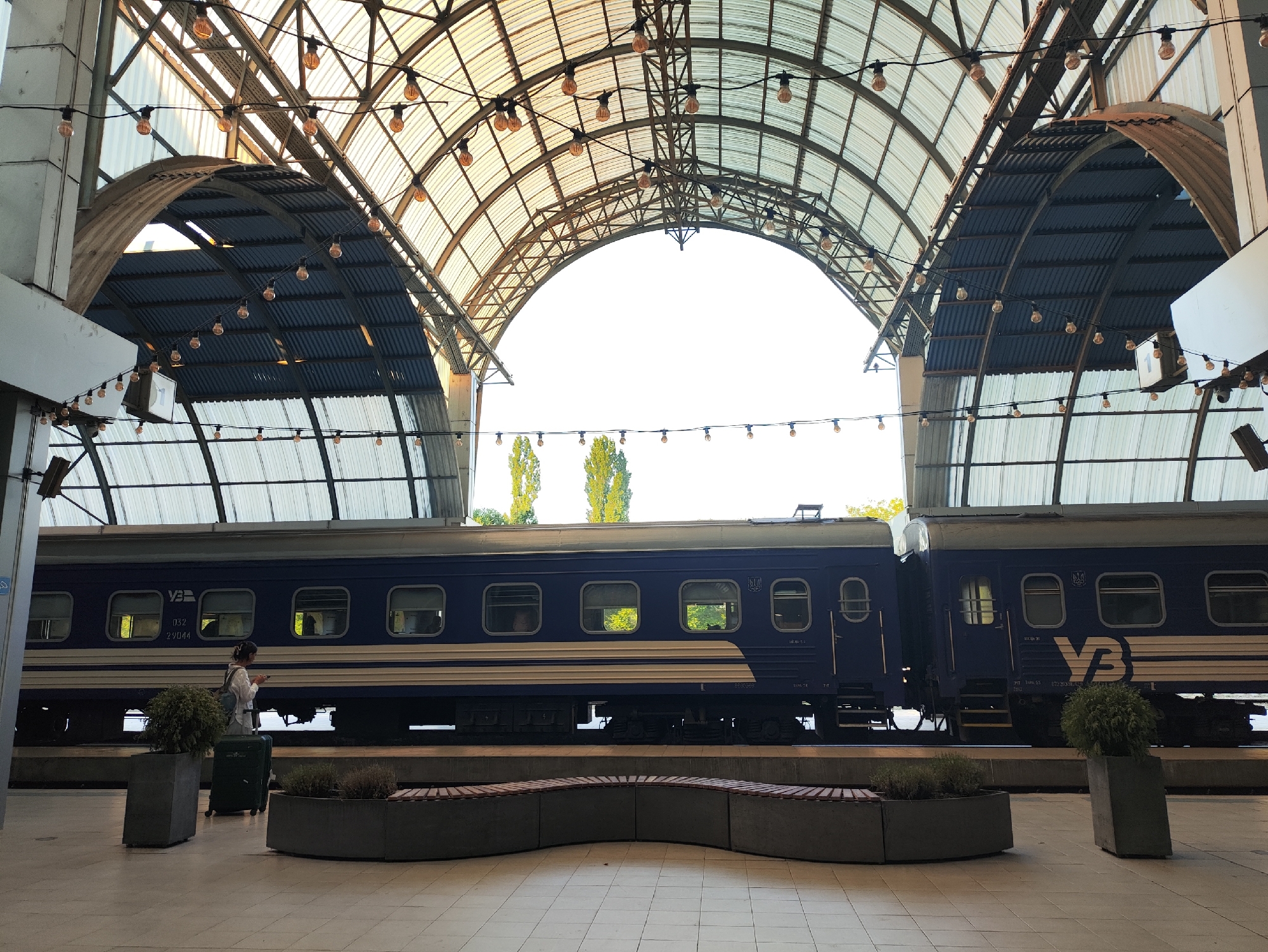 This is an image of a train at a station. The train is dark blue with a white stripe. There are several windows visible on the side of the train. A woman with a backpack and a suitcase is standing on the platform next to the train. She is looking at her phone. The platform is paved with light-colored tiles. There are two planters with green bushes on the platform, and a curved bench. Above the platform is a large, arched roof structure made of metal and translucent panels, with many strings of lights hanging from it. In the background, beyond the train, are some tall trees and a bright sky. This scene is likely at the Chișinău Railway Station in Chișinău, Moldova, given the distinctive architectural style of the station's roof and the presence of a train consistent with those used in Eastern Europe.