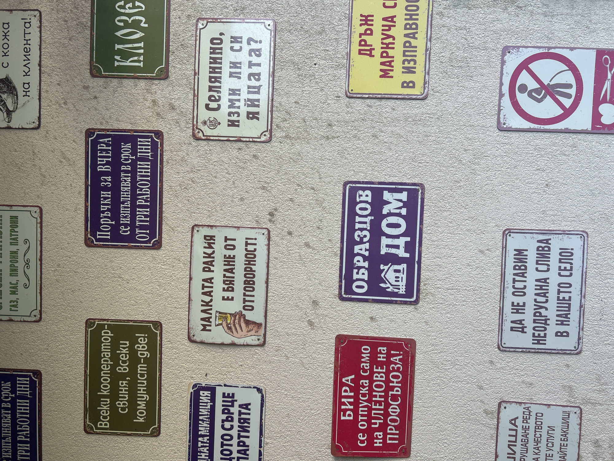 The image shows a collection of small, weathered, vintage-style signs mounted on a textured surface, possibly a wall. The signs feature text in Bulgarian and various illustrations, alluding to everyday life, humor, and satirical commentary on aspects of Bulgarian culture and society, possibly during the communist era. The signs' topics range from practical instructions and product advertisements to jokes about rural life, bureaucracy, and social dynamics. The signs' worn appearance and design suggest that they are intended to evoke a sense of nostalgia and amusement. The location of Konska, Bulgaria is not depicted in the image.