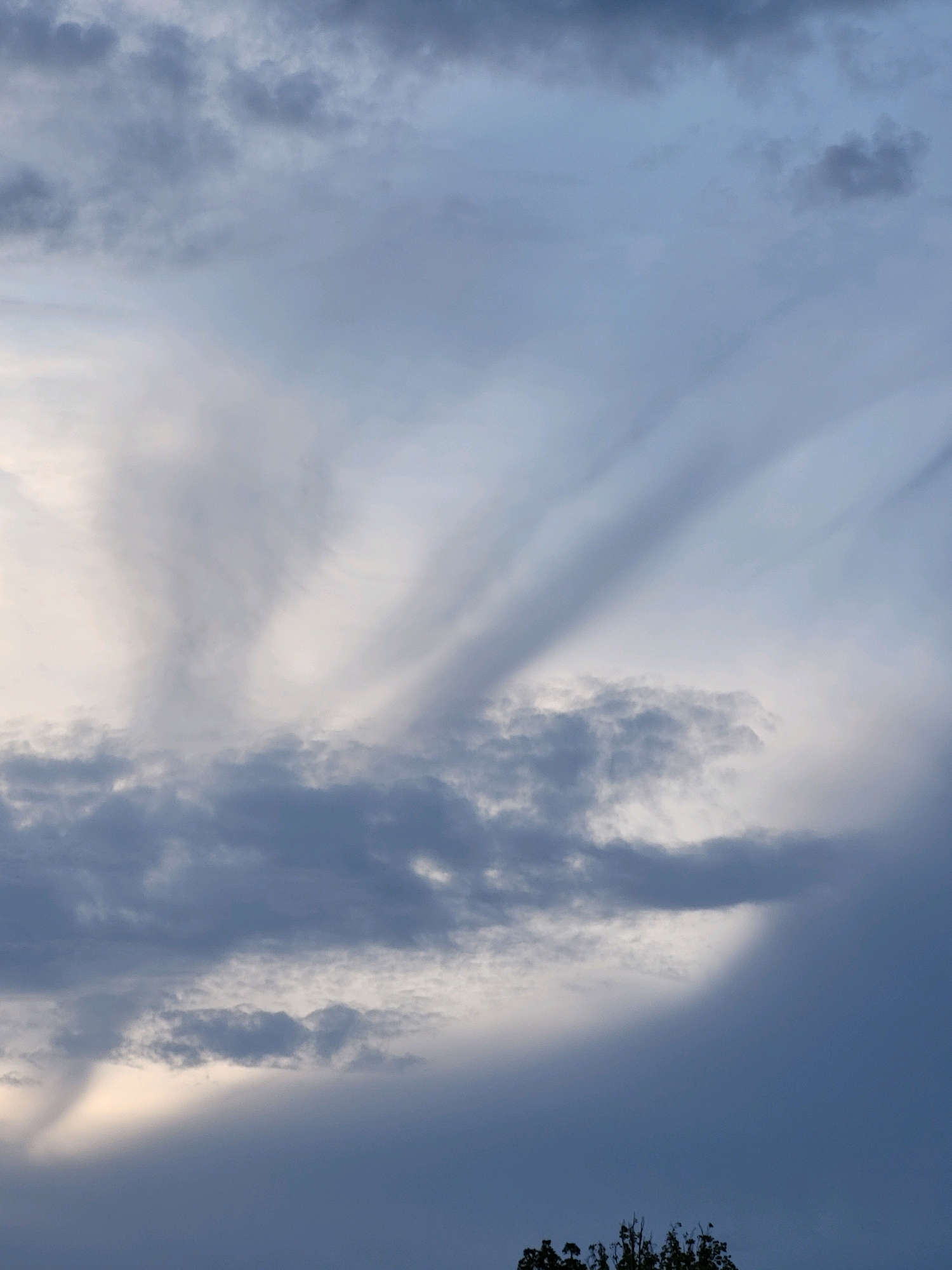 The image shows a cloudy sky with a hint of sunlight breaking through the clouds. The clouds are varied in texture and density, with some appearing wispy and others more solid. The overall color palette is dominated by shades of blue and gray, with a soft yellow glow emanating from the area where the sun is partially visible. The lower portion of the image includes the dark silhouette of treetops, suggesting an outdoor scene. The location is specified as Ozolnieki, Latvia.
