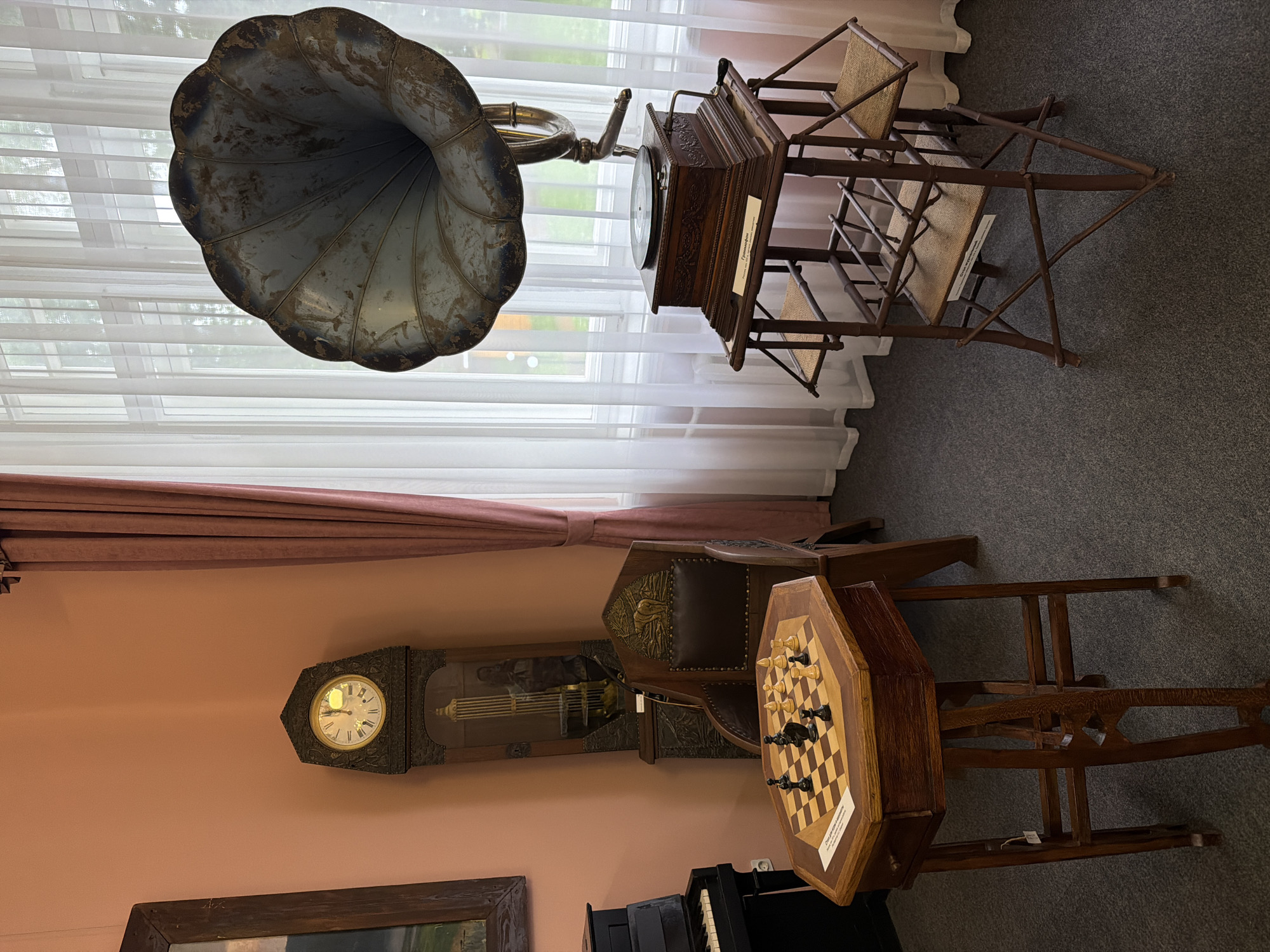 This media file depicts an indoor scene showcasing antique items. The main subjects are a gramophone with a large horn, a grandfather clock, a chessboard with pieces set up, and parts of a piano. The setting appears to be a room with a large window covered by sheer white curtains and a pink wall. There are also wooden chairs and a table. The floor is covered with dark grey carpet.
The gramophone is prominently featured in the upper left, its metallic horn pointing upwards and to the left. Next to it, a wooden cabinet with a record player is visible, supported by a rustic wooden stand with woven seats. A label is attached to the stand.
In the lower section, a grandfather clock with an ornate dial and pendulum is mounted on the pink wall. To its right, an octagonal wooden table holds a chessboard with black and white pieces in mid-game. Behind the chessboard, a leather-upholstered chair is visible. A portion of a piano keyboard is visible in the bottom center.
The lighting suggests daytime, with light streaming through the window. There are no people visible, and the scene is static, suggesting a display or a preserved historical room. The overall impression is one of nostalgia and historical preservation. No specific weather conditions can be inferred. The location context is Sarapul, Russia, but no explicit visual cues in the image directly confirm this. There is text on a label attached to the wooden stand of the gramophone, and also on a label on the chessboard, but the text is not legible enough to be transcribed.
