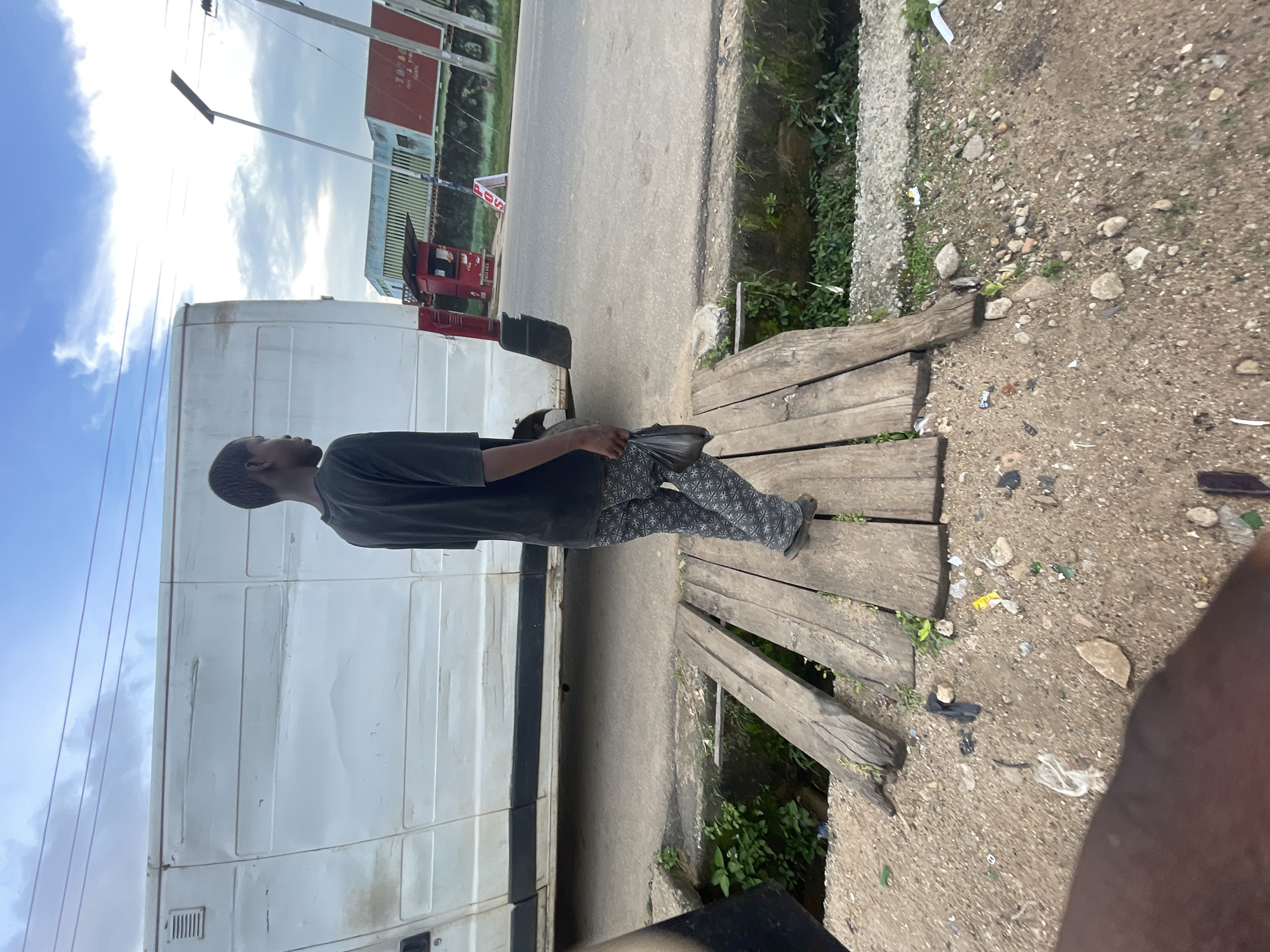 A young Black male, dressed in a dark t-shirt and patterned pants, walks across a makeshift wooden bridge. He carries a dark plastic bag in his right hand. The bridge spans a narrow, overgrown ditch filled with green vegetation. Behind him, a large white vehicle, possibly a van or bus, is parked. The scene appears to be a roadside in Jos, Nigeria, with a paved road and buildings visible in the background. The sky is overcast with some visible clouds and power lines. The ground is rough, with scattered debris and gravel. The overall impression is one of a common, everyday scene in an urban or semi-urban environment.