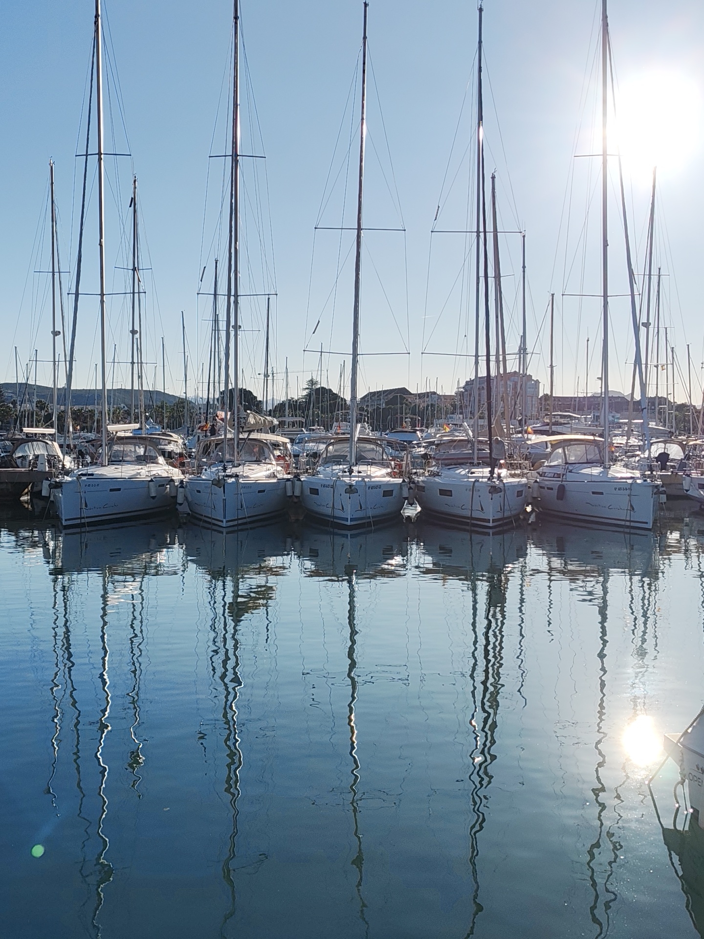 This media file captures a serene and sun-drenched marina scene in Dénia, Spain. Numerous white-hulled sailboats, mostly of the "Valiant Capt'n" model, are moored closely together, forming neat rows. Their tall, slender masts and intricate rigging dominate the upper half of the image, crisscrossing against a clear, bright blue sky.
The boats are tightly packed, with their sterns facing the viewer. Visible registration numbers include "6a BA-3-12-22", "6a BA-3-13-17", and "6a BA-3-35-13" on various vessels. The calm, undisturbed water of the marina reflects the boats' masts, hulls, and rigging with remarkable clarity, creating a mesmerizing pattern of vertical lines and shimmering light.
The sun, positioned off-center to the right, casts a brilliant glare on the water and through the rigging, indicating either early morning or late afternoon. In the background, behind the moored boats, a coastline with lush greenery, low-rise buildings, and distant hills or mountains is visible under the bright sunlight. The overall atmosphere is peaceful and bright, showcasing a typical sunny day in a Mediterranean port.