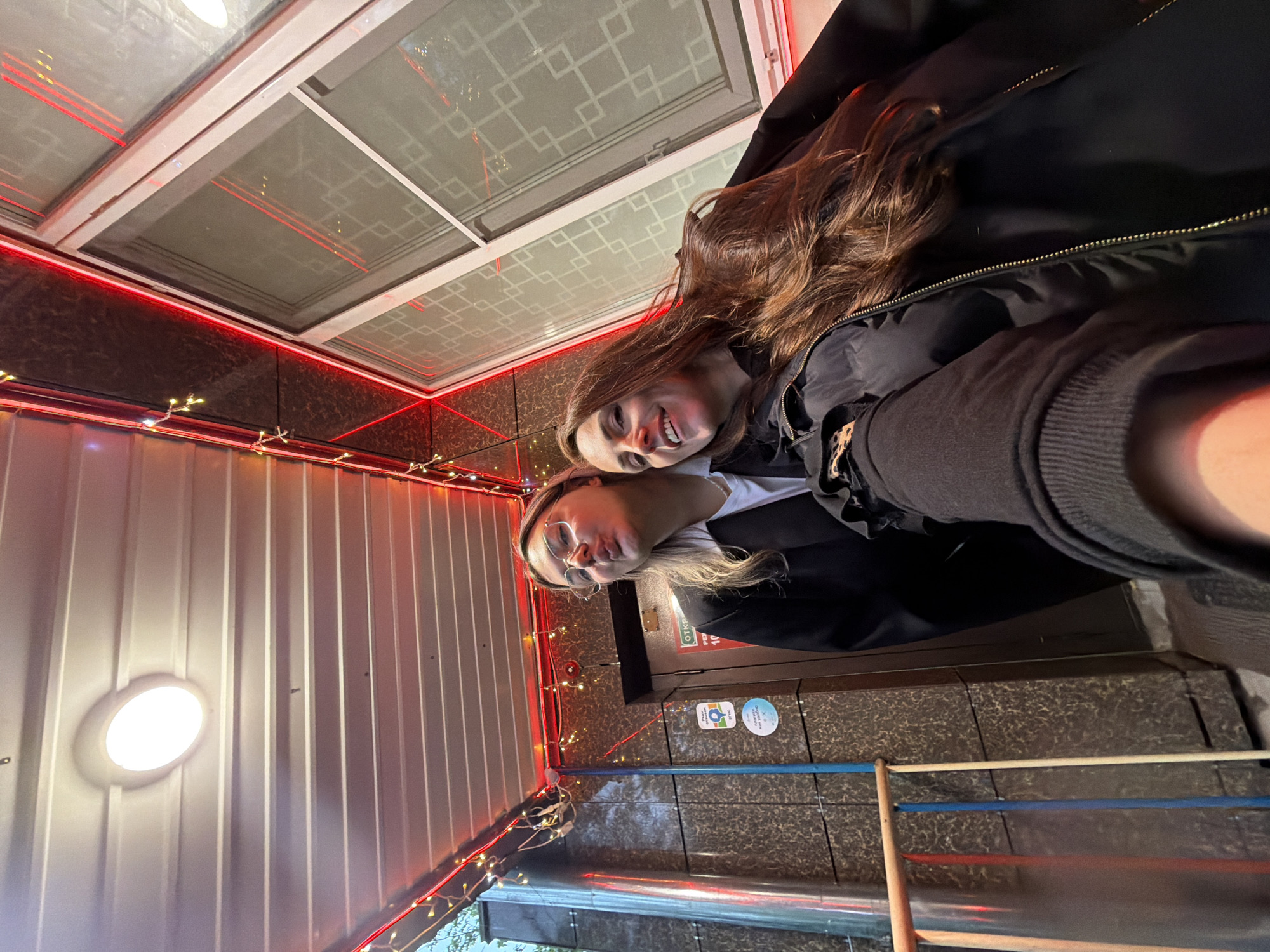 Two young women pose for a selfie in what appears to be a covered outdoor area or an entranceway, likely in the evening or at night given the artificial lighting. The image is captured from a slightly upward angle, giving a dynamic, casual feel.
The woman on the right has long, wavy brown hair, a bright smile, and is wearing a dark, possibly padded, jacket. She is positioned slightly behind and above the other woman. The woman on the left has blonde or light brown hair, wears glasses, and is making a playful "puckered lips" expression. She is dressed in a black jacket over a white top. Both women exhibit happy and friendly emotions.
The setting features an overhead structure with glass panels, reflecting red LED strip lights that run along the edges of the ceiling and walls. These glass panels have a distinctive geometric pattern. Additional small, warm-toned string lights (fairy lights) are draped around the structure. One wall to the left consists of corrugated metal with a single round, bright light fixture. To the right, another wall is covered in dark, textured tiles or panels. A door, partially visible behind the blonde woman, has text in Russian, which reads "ОТКРЫТО" (OTKRYTO), meaning "OPEN." Below this, two circular stickers with symbols are affixed to the wall. In the lower right of the frame, several colorful pipes (red, blue, beige) are visible, possibly part of a decorative railing. The overall atmosphere suggests a casual, perhaps commercial, outdoor space in Birobidzhan, Russia.