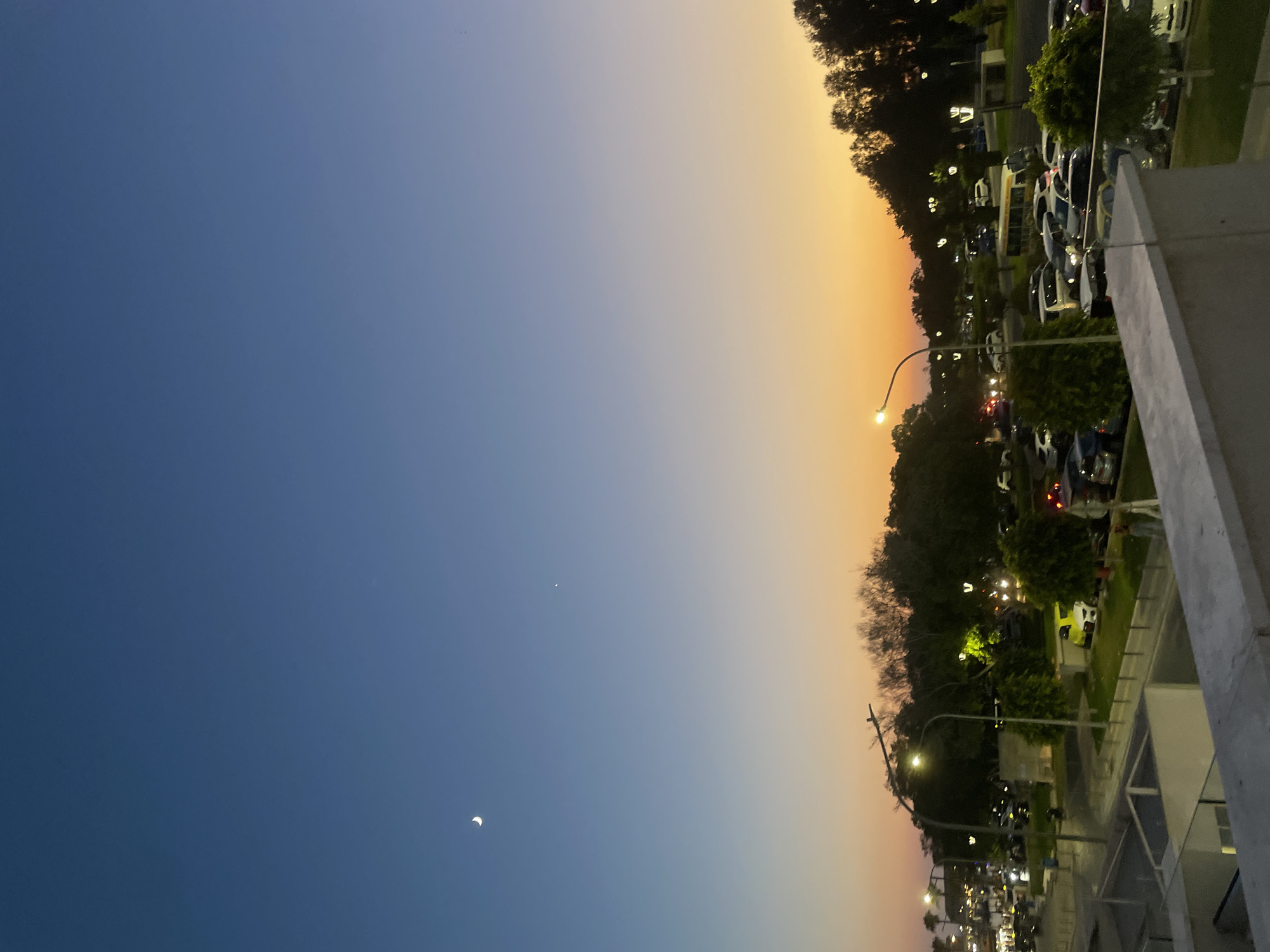 The image depicts a twilight scene in Paphos Municipality, Cyprus. The sky exhibits a gradient of colors, transitioning from a deep blue at the top to a warm orange and yellow near the horizon, indicating sunset or sunrise. A crescent moon is visible in the upper left portion of the sky, accompanied by a small, bright celestial body to its right.
Below the sky, a line of trees forms a dark silhouette against the vibrant sky, with some of the trees illuminated by streetlights. The ground level shows a parking lot filled with numerous cars of various colors and models. Streetlights are strategically placed throughout the area, casting a soft glow on the surroundings. A bus is also visible among the parked vehicles.
The foreground of the image is occupied by a concrete balcony or ledge, suggesting an elevated viewpoint. The overall impression is that of an urban or semi-urban environment during the transitional period of twilight. The presence of cars and streetlights indicates human activity and infrastructure. No people are clearly discernible in the image, though the presence of a populated parking lot implies their proximity. The environment appears to be a public space, possibly a park or a commercial area with ample parking facilities.