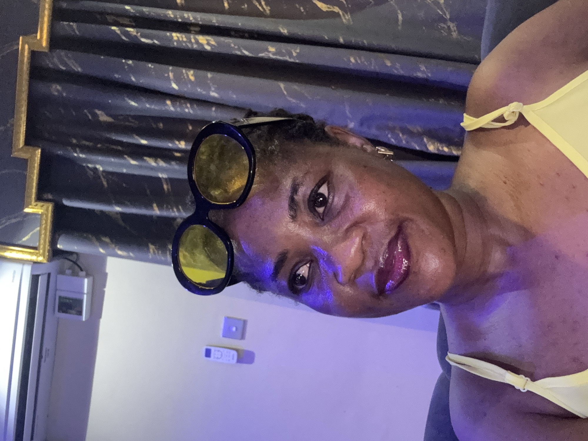 The image shows a close-up of a Black woman wearing yellow sunglasses on her head and a yellow top. She has dark skin, dark hair, and is wearing lip gloss. She is looking to her left. In the background, there is a dark blue curtain with gold accents and a white air conditioning unit mounted on a light-colored wall. There are also two white electrical outlets or switches on the wall. The lighting in the scene is predominantly purple. The image appears to be taken indoors, possibly in a residential setting in Ajara, Nigeria.