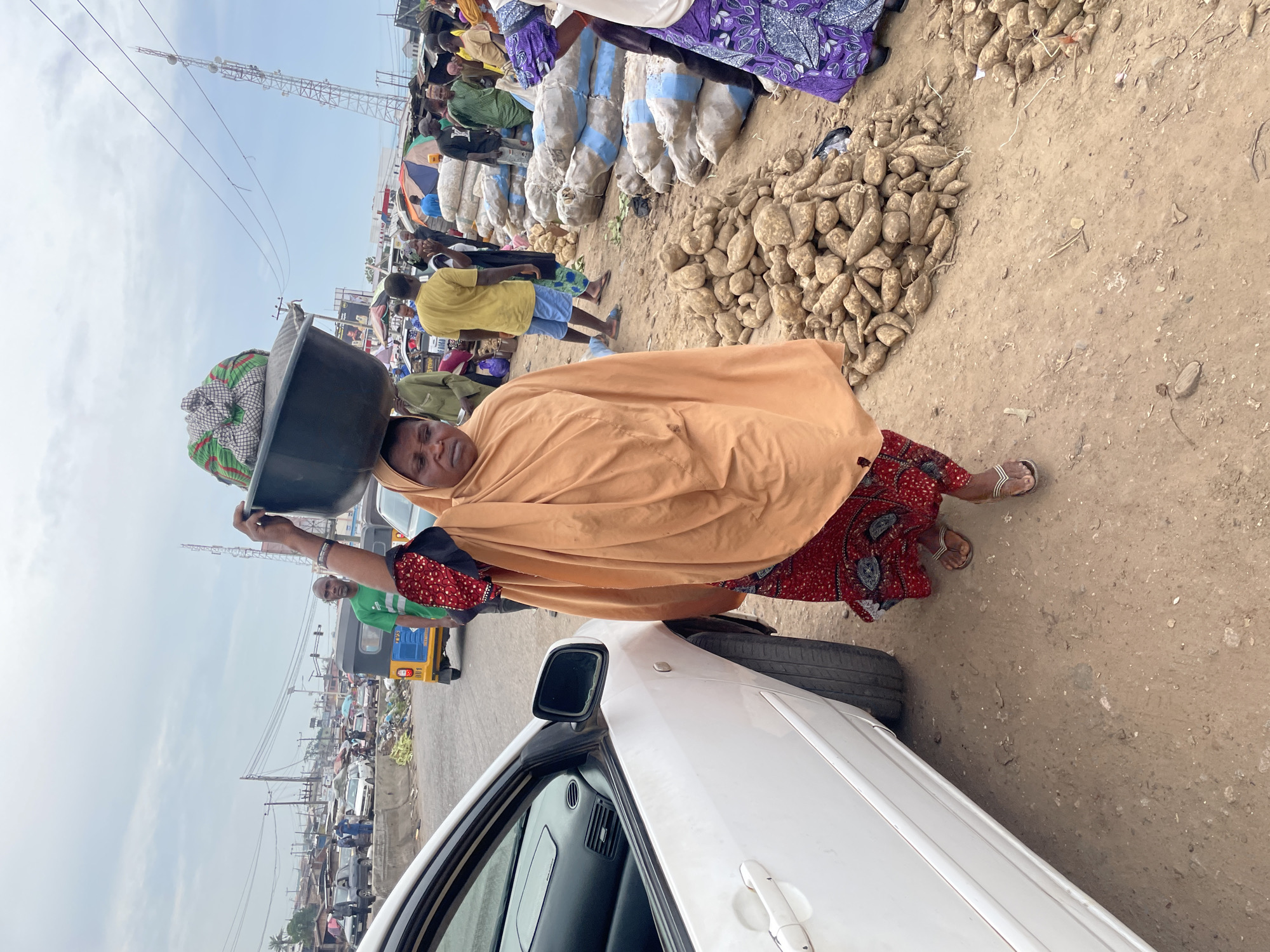 In a dusty outdoor market in Lokoja, Nigeria, a woman walks with a large black basin filled with goods balanced on her head. She is wearing a vibrant red and black patterned skirt and an orange headscarf and shawl. To her left, the rear of a white car is visible. In the background, a busy street scene unfolds with various vehicles, including a yellow auto-rickshaw, and other market-goers are seen browsing stalls. Several large sacks of produce, likely yams, are piled on the ground to the right of the woman, with more yams in a mound in the foreground. The sky is overcast.