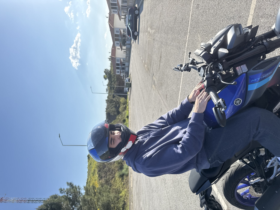 The image depicts a male individual seated on a blue and black Yamaha MT-125 motorcycle in an asphalt parking lot under a clear blue sky in Mirandela, Portugal. The subject wears a dark blue hoodie, dark blue jeans, and a multi-colored full-face motorcycle helmet with the visor partially open, revealing his face. His hands rest on the motorcycle's fuel tank. In the background to the left, a green hillside with some yellow vegetation is visible, topped by a red and white communication tower and a tree. To the right, a residential area features several two-story houses with light facades and red-tiled roofs, fronting a paved street where a black hatchback car is parked. Multiple streetlights are present in the background. White parking lines are visible on the asphalt surface. The scene is illuminated by bright sunlight.