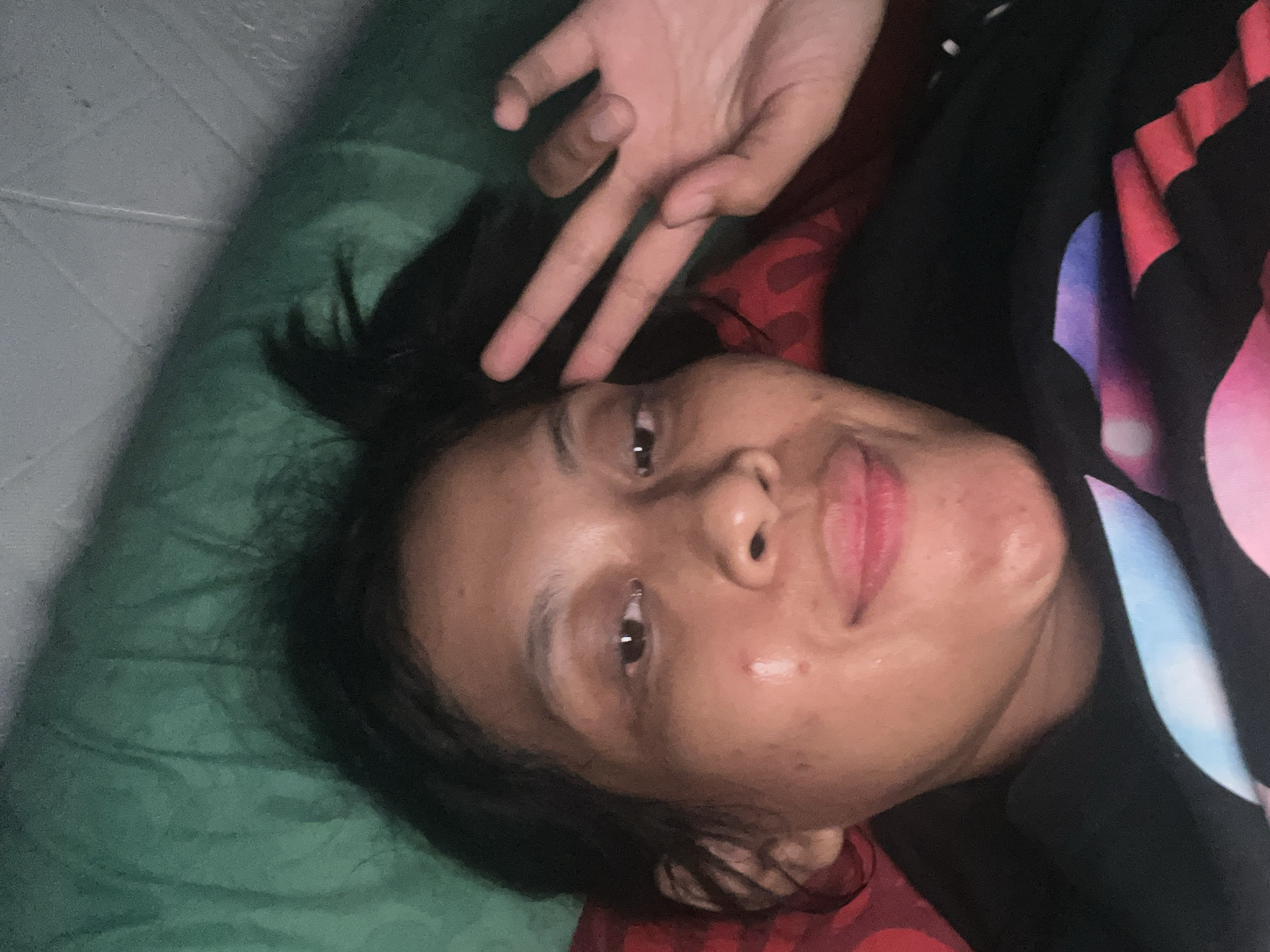 The image shows a close-up, selfie-style portrait of a young woman lying down. She is looking directly at the camera with a slight smile and has her right hand raised, forming a peace sign. Her hair is dark and pulled back loosely. She is wearing a dark colored t-shirt with a colorful, abstract graphic design. The background appears to be a green surface, possibly a blanket or pillow, and a red textured object, possibly another pillow. The lighting suggests it may be indoors or at night. The specific location in Kecamatan Pati, Indonesia, cannot be determined from the visual information provided in the image.
