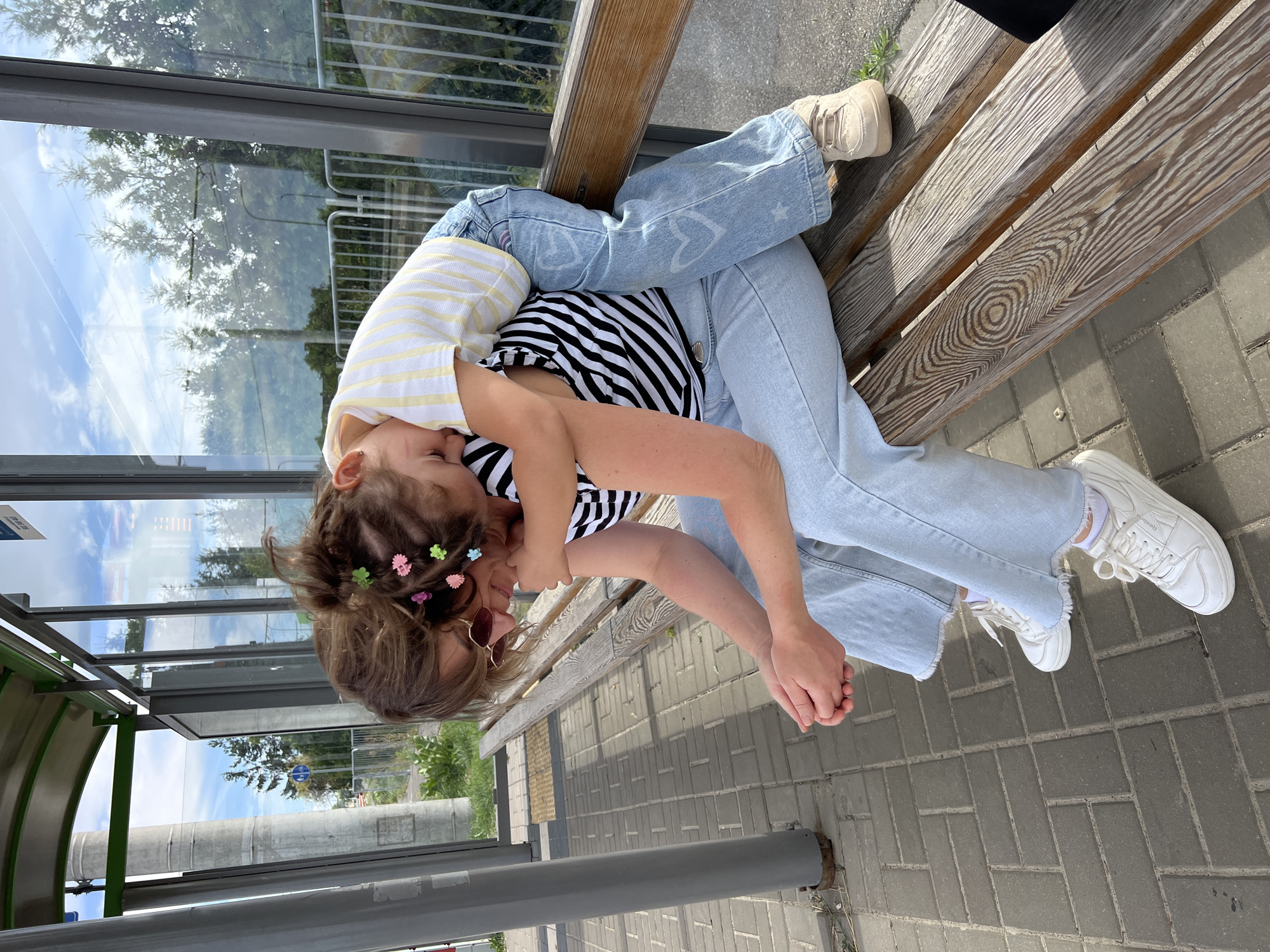 A woman and a child are sitting on a wooden bench at a bus stop in Lublin, Poland. The woman is wearing a black and white striped t-shirt, light blue jeans, and white sneakers. She has sunglasses on and her hands are clasped in her lap. The child is sitting on the woman's lap, facing away from the camera, and is wearing a yellow and white striped t-shirt and light blue jeans with white hearts and stars printed on them. The child has her arm around the woman's neck. The bus stop has a glass enclosure and a green metal roof. The ground is paved with gray bricks. The background shows trees and a blue sky with clouds.
