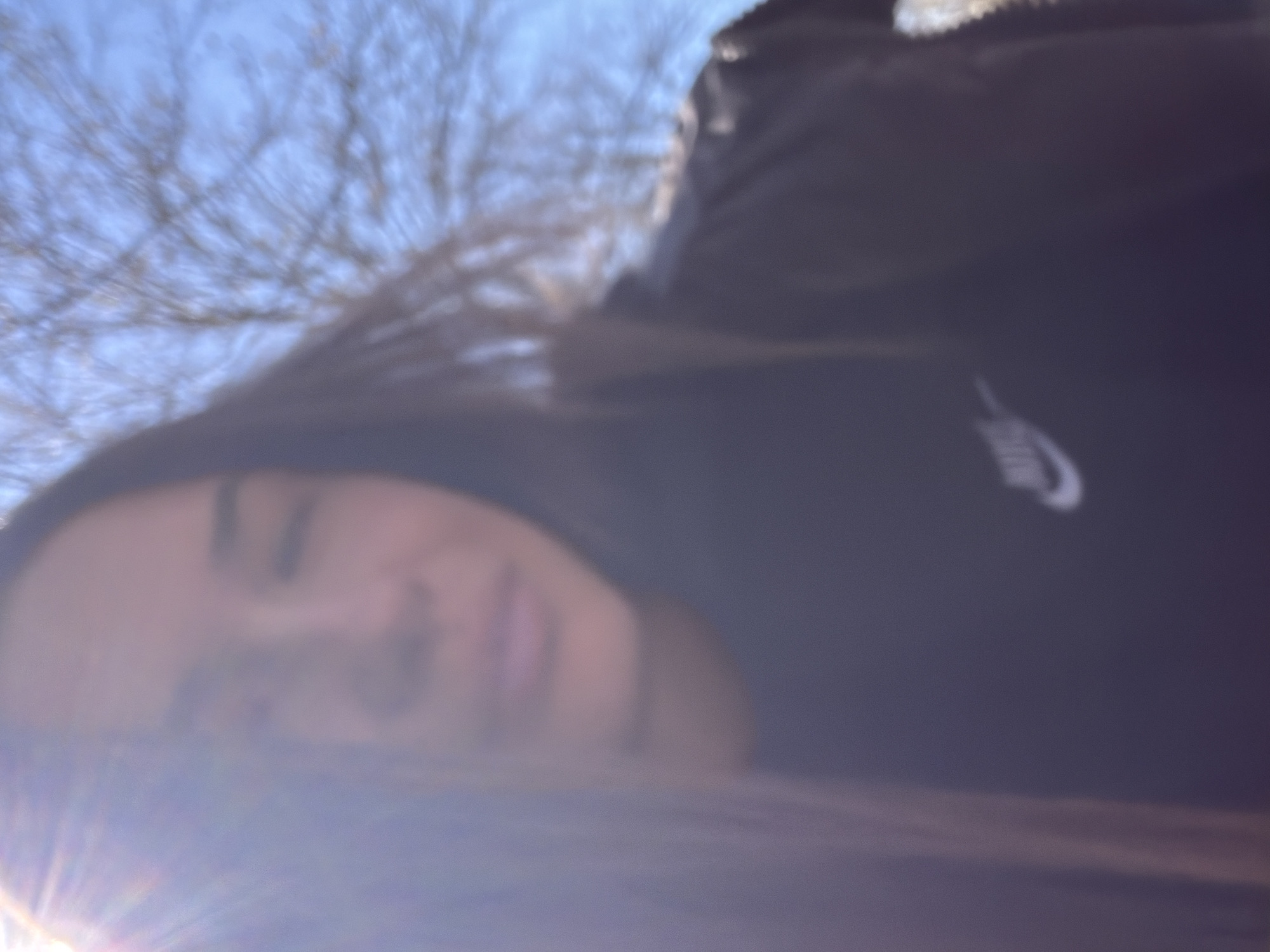 A blurred photograph shows a person with dark hair and a dark jacket with a white Nike logo. The background includes leafless tree branches against a light blue sky. The location cannot be determined from the image.