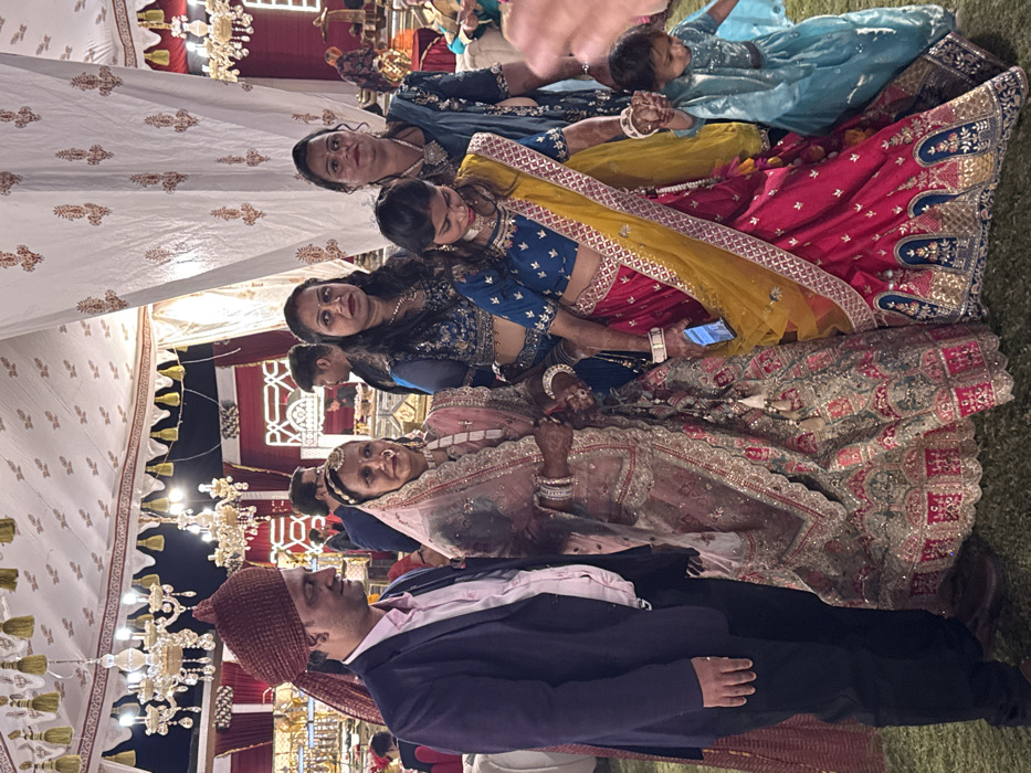 A group of people poses for a photo at what appears to be a wedding reception. The setting is under a white tent adorned with chandeliers. A man in a dark suit and turban stands in the foreground. Behind him, several women are dressed in traditional Indian attire. One woman holds a baby in a light blue outfit. The other women wear colorful saris with intricate embroidery and jewelry. One woman holds a smartphone in her hand.