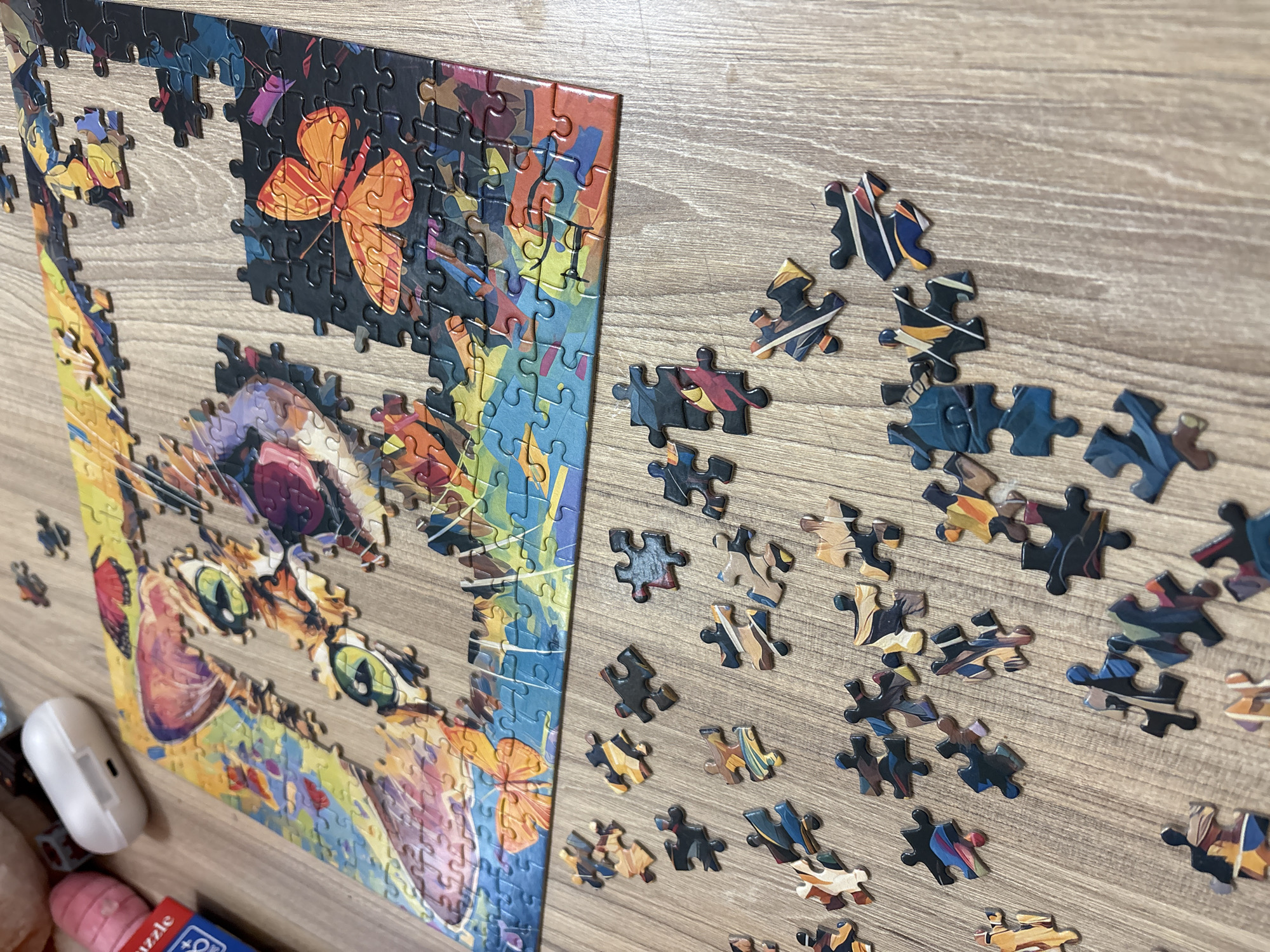 The image shows a partially completed jigsaw puzzle on a wooden surface. The puzzle depicts a colorful cat with green eyes and an orange butterfly in the upper portion. Many loose pieces, primarily dark-colored with hints of blue, orange, and yellow, are scattered beside the assembled section. Near the bottom left corner are some blurred items, including what appears to be a white earbud case and part of the puzzle box. The location, Panagyurishte, Bulgaria, is not discernable from the given image. There are no people visible.