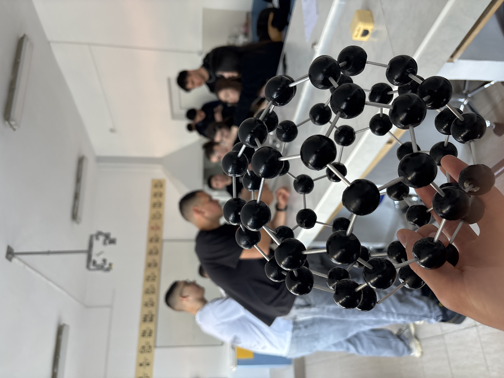 A medium, indoor shot presents a molecular model of Buckminsterfullerene, constructed of black spherical atoms joined by metallic rods, held by a right hand in the foreground. The background shows a classroom or lab setting, with students seated at tables. Most of the students are male, with dark hair, wearing dark attire. The environment includes white walls with overhead lighting fixtures, possibly fluorescent. A scale is visible on the wall. The surfaces appear clean and the setting is academic. The location is not apparent from the image and there is no information to indicate Zlatograd, Bulgaria.