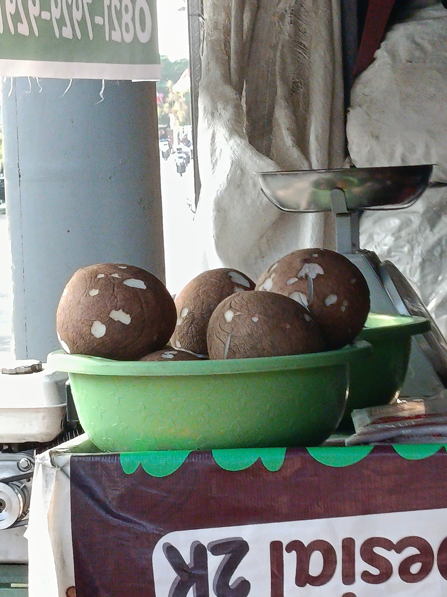 The image depicts a street-side stall, likely in Palembang, Indonesia, where coconuts are being sold. In the foreground, a green plastic bowl holds several whole coconuts, each with its distinctive brown husk and patches of white where the husk has been partially removed. To the left, a portion of a white and grey motorized unit, possibly part of a generator or a machine for processing coconuts, is visible. Above the coconuts, a metal weighing scale with a round pan is positioned. In the background, a banner with green and white lettering, partially obscured, suggests pricing or branding. Further back, a white cloth hangs, creating a backdrop. Outside the stall, a blurry view shows traffic, including motorcycles, indicating an urban environment. The lighting suggests it is daytime, with sunlight casting shadows.