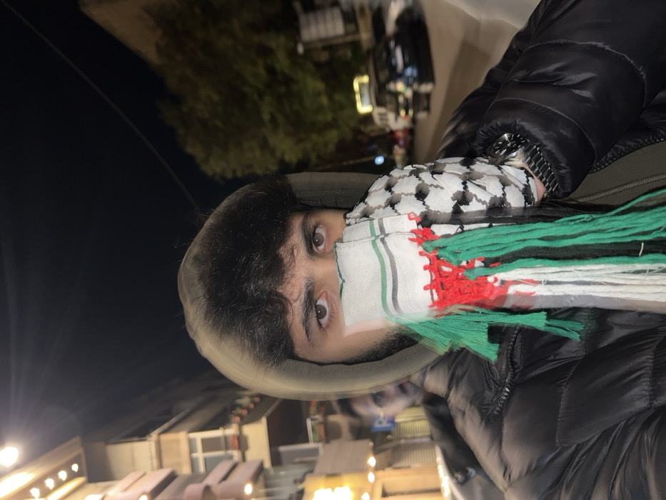 A person wearing a black puffer jacket and a hooded sweatshirt is partially covered by a kaffiyeh, with the bottom half featuring green, white, and red stripes with tassels. Only the person's eyes and forehead are visible. The background shows a dark sky, some foliage, and indistinct buildings illuminated by streetlights at night. The image is rotated, causing the background to appear vertically along the left side.