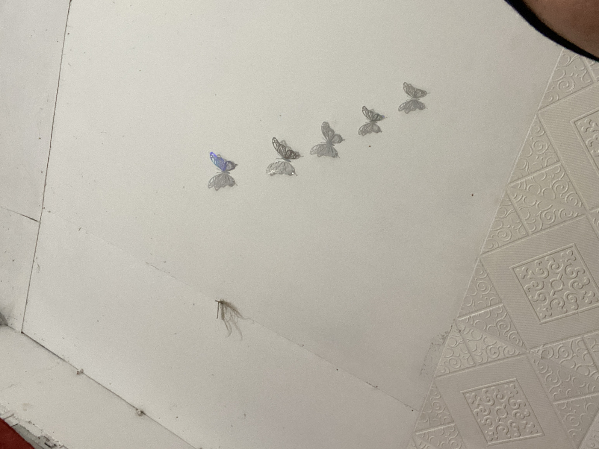 A dead mosquito lies on a white, textured surface, likely a ceiling or wall, in Banjarbaru, Indonesia. The mosquito is positioned on a diagonal seam, with its legs splayed out. Six metallic butterfly stickers are arranged in a diagonal line across the white surface, some appearing to have a holographic sheen. The lower portion of the image shows a section of patterned tiles. The scene is lit from above, casting subtle shadows.