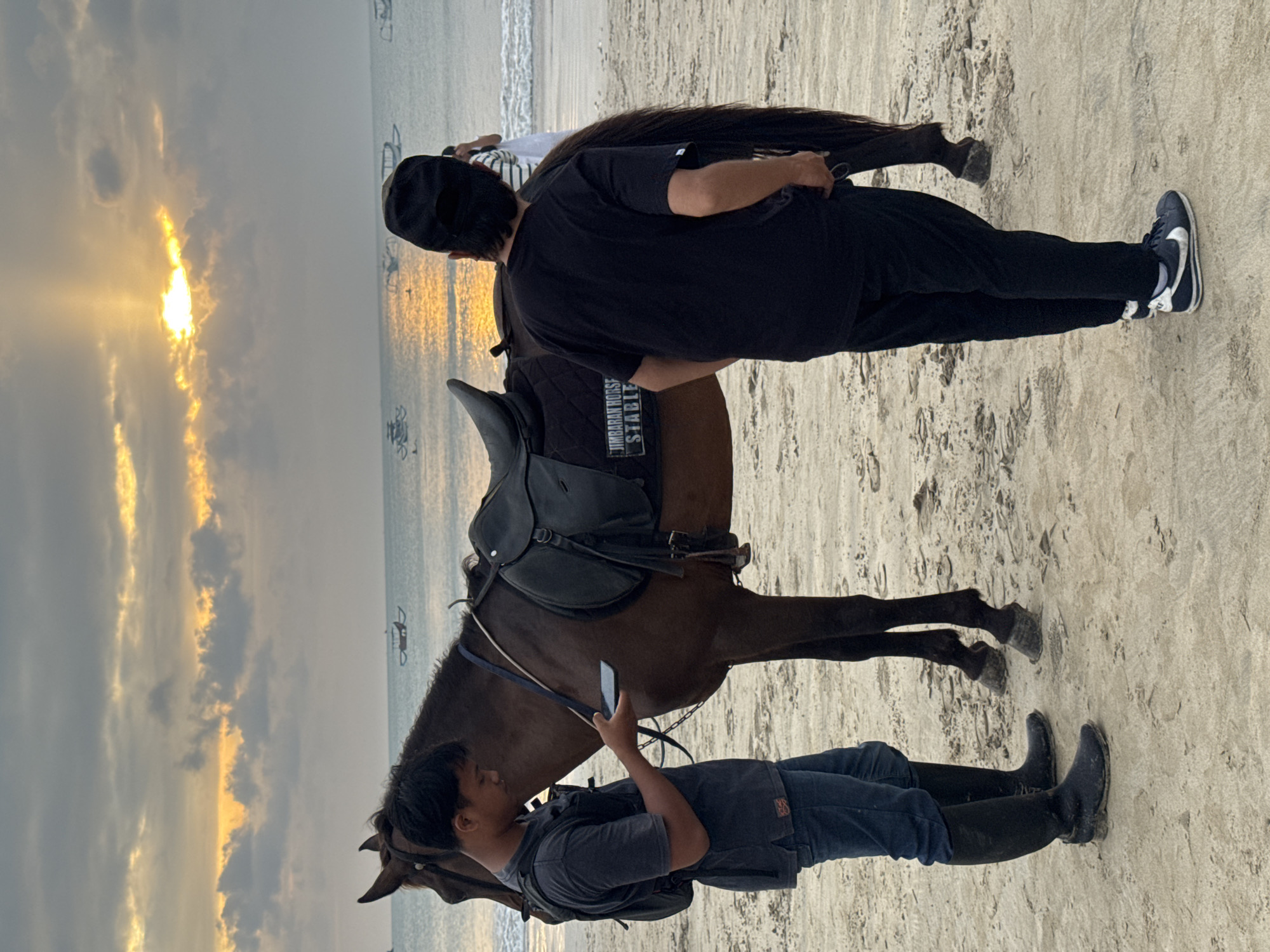 An overcast sunset casts a warm glow over Jimbaran Beach in Kuta Selatan, Indonesia. Two men stand on the sandy shore next to a brown horse. The man on the left, wearing a dark t-shirt, blue jeans, and tall black boots, is looking at his phone. The man on the right, dressed in black pants, a black t-shirt, and a black baseball cap, stands with his back to the camera. The horse is saddled with a black saddle, and a saddle pad with the text "JIMBARAN HORSE STABLE" is visible. In the distance, the ocean is dotted with traditional Balinese fishing boats, and the sun's rays break through the clouds, reflecting on the water. The sand is light brown and textured with footprints.
