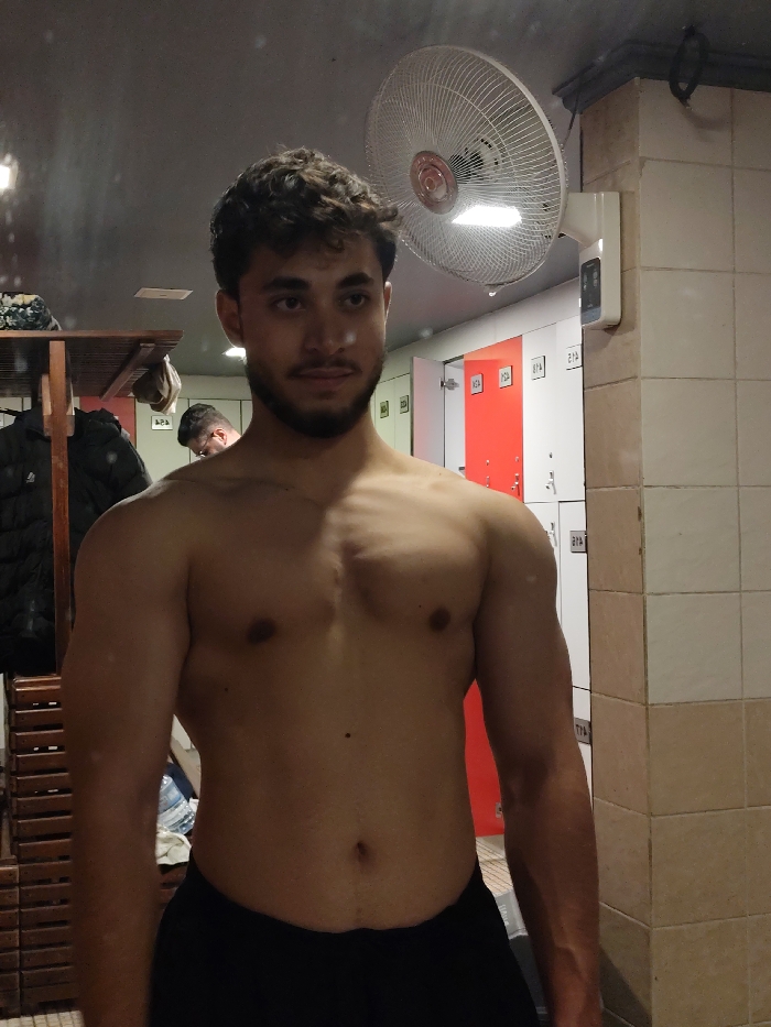 A shirtless man with curly hair and a beard stands in a locker room, posing in front of a mirror. He is wearing black athletic shorts. In the background, there are lockers, some of which are red and white. A fan is mounted on the wall above the lockers. Another person is partially visible in the reflection of the mirror, in the background of the image.