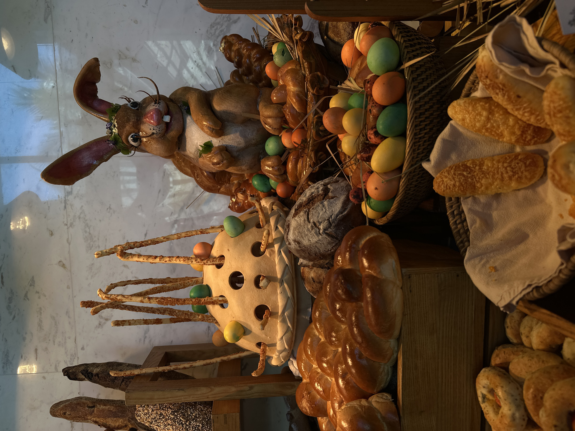 The image depicts a decorative display of Easter-themed items, likely set up in a commercial establishment in Nusa Dua, Indonesia.
Foreground:
A large, cartoonish rabbit figure with a floral wreath around its head is prominently displayed. A decorative object with holes filled with colored eggs and bread sticks. Several loaves of braided bread and various other breads are arranged.
Background:
Baskets filled with colorful Easter eggs and various bread types. The overall arrangement is intended to be festive and celebratory, targeting customers of the establishment.
Setting:
The image was likely taken indoors, indicated by the lighting and the marble-like tile floor. The scene suggests a bakery or a restaurant setting, and the decoration clearly targets Easter festivities.
