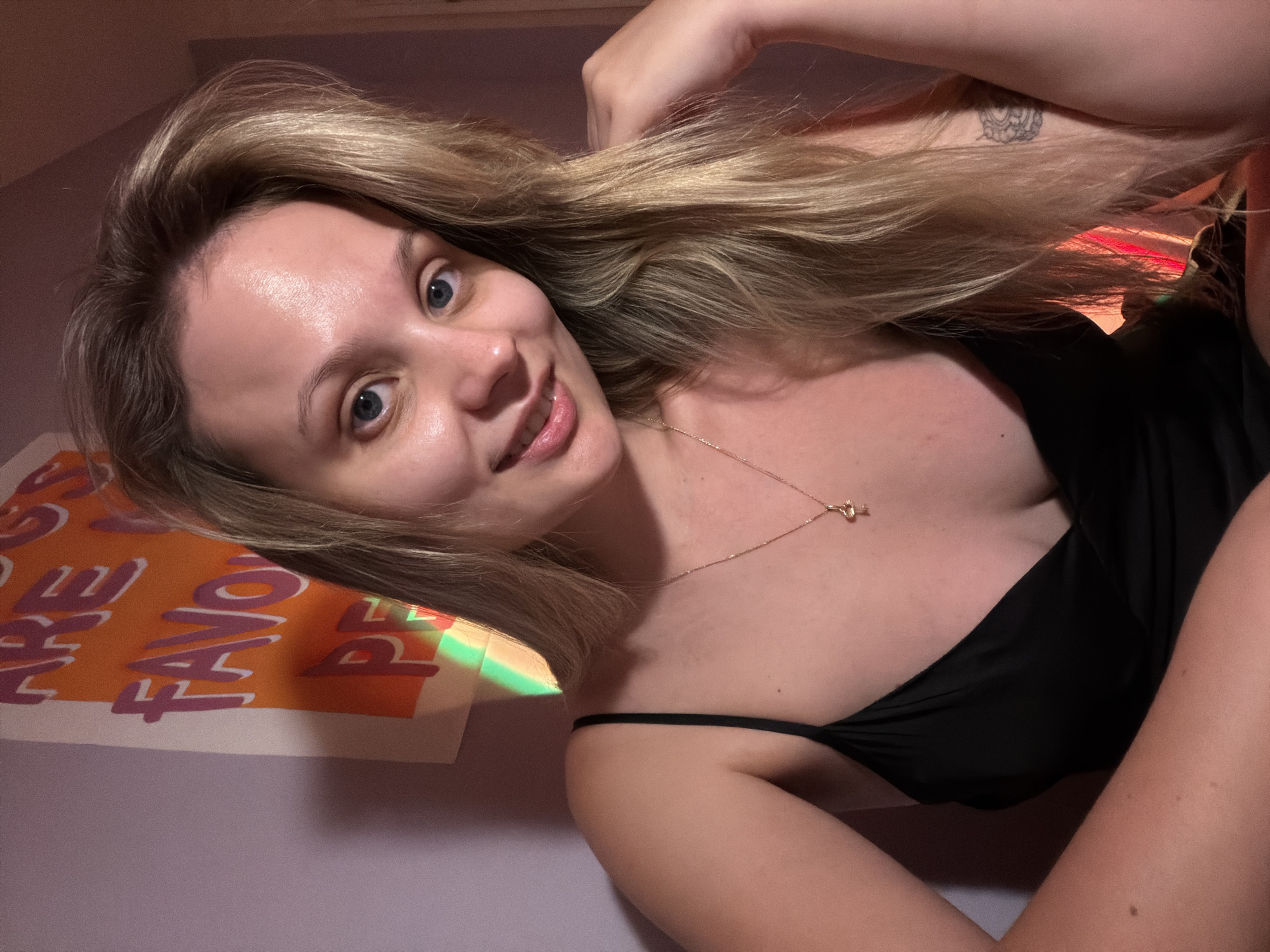 A woman with blonde hair and blue eyes is lying down, looking at the camera. She is wearing a black top and a gold necklace. She has a tattoo on her left arm. Behind her is a poster with the words "YOU ARE THE FAVO." The background appears to be a room with a lavender wall. The location is not discernible as Tel Aviv, Israel.