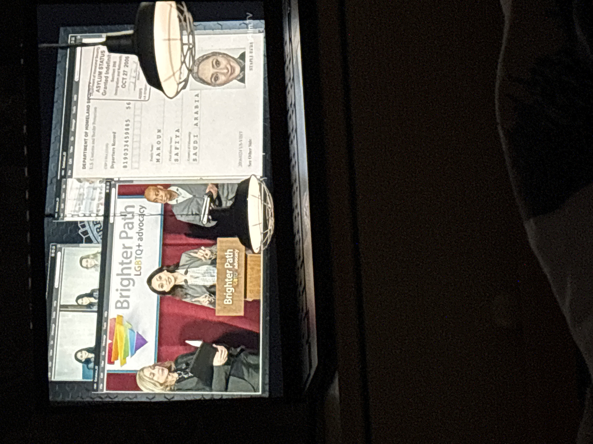 The image shows a display featuring several elements. At the top, there is what appears to be a Department of Homeland Security Departure Record granting asylum status to Safiya Maroun, originally from Saudi Arabia, dated October 27, 2006. Below that is a promotional display titled "Brighter Path LGBTQ+ Advocacy," depicting various people. The environment is dark, and the image appears to be taken indoors, possibly within a museum or exhibit setting. The city and country are not discernible from the image.