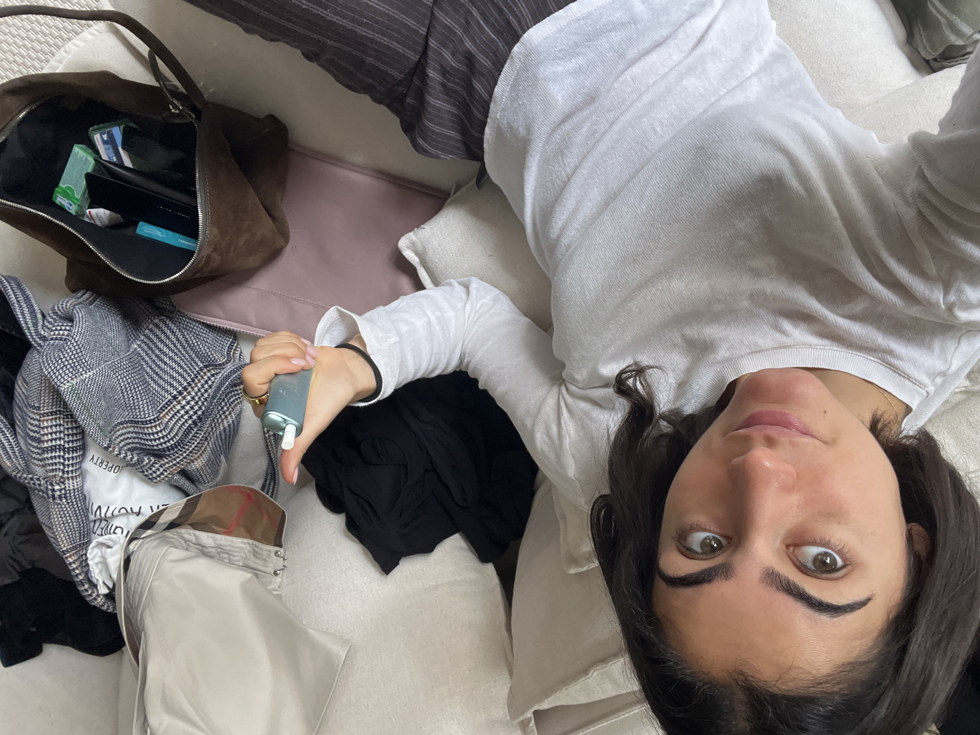 The image shows a woman lying on a light-colored sofa, surrounded by various objects. To her left, a brown suede handbag sits next to a light pink bag. Clothes are scattered around her, including a plaid jacket, a white t-shirt, and a black garment. The woman is holding a light blue object in her hand. The location is not specified, so it is impossible to verify if it is Paris, France.