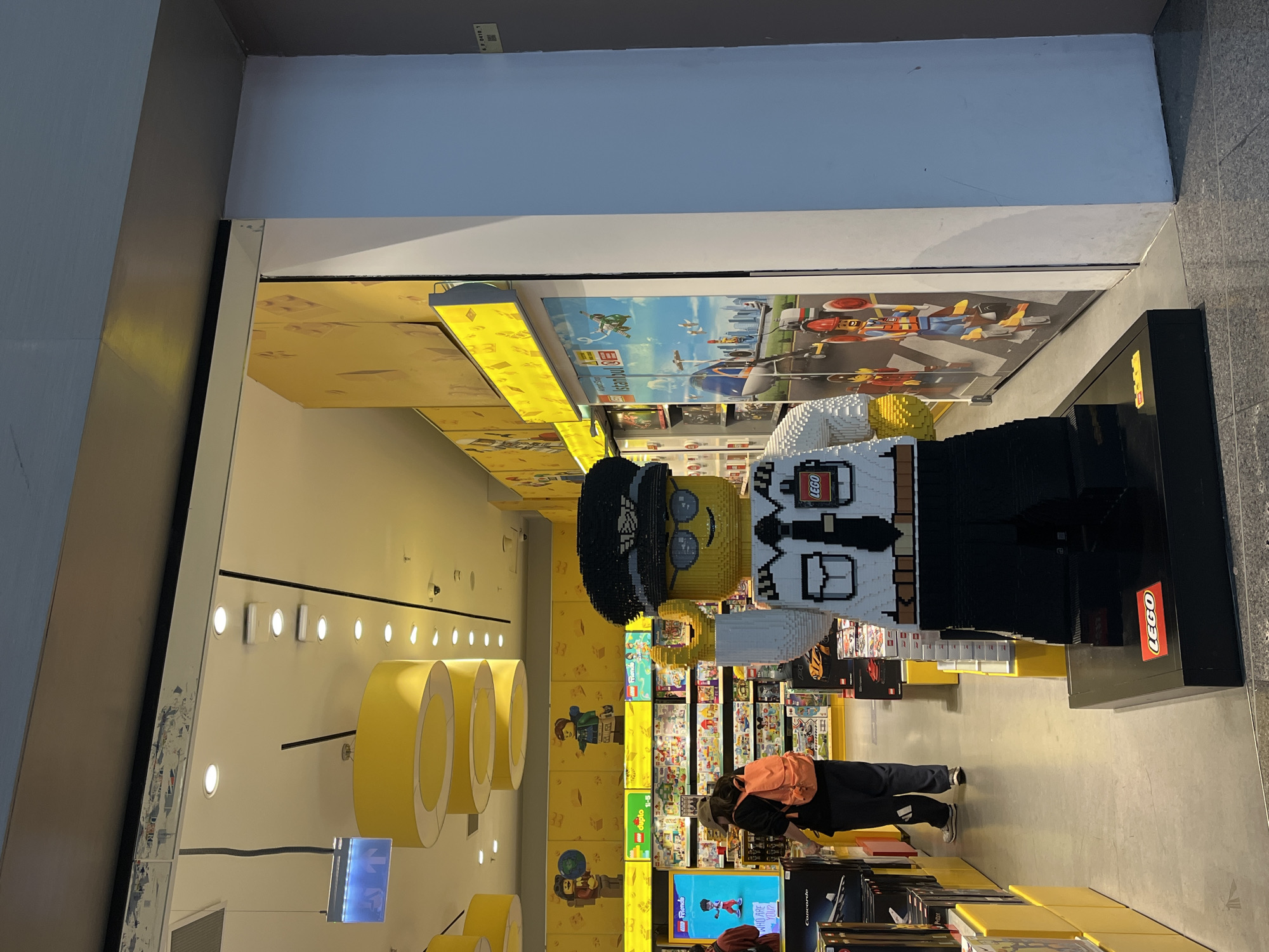 The image depicts the entrance to a LEGO retail store, identified by an internal poster as "Airport Shop Istanbul," placing the location in Istanbul, Turkey. A life-sized LEGO pilot figure, constructed from yellow, white, and black bricks, stands prominently on a black base with a red LEGO logo at the right side of the entrance. The figure wears a white shirt, black tie, black epaulets, a black pilot's hat with a white winged emblem, and sunglasses; its right arm is slightly raised.
Inside the store, yellow walls are adorned with large LEGO character decals. The ceiling is equipped with numerous recessed circular lights and three large yellow cylindrical light fixtures. The floor consists of light grey tiles. One individual, viewed from behind, wears a black top, dark grey sweatpants with white stripes, grey sneakers, an orange backpack, and a beige baseball cap, and is observed interacting with merchandise on shelves to the left. A second individual, wearing a red top, is partially visible on the far left. The shelves are stocked with various LEGO sets, with overhead signage indicating categories such as "LEGO Duplo 1-5" and "LEGO Friends." A vertical digital display on the left exhibits a LEGO figure and text. A large wall poster to the right of the entrance illustrates an airport scene populated by LEGO elements and characters. A faded blue exit sign hangs from the ceiling on the upper left interior. The entrance structure is light grey, and the overhead ceiling trim exhibits surface wear.