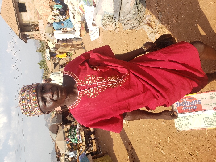An outdoor scene in a market area of Adonkolo, Nigeria, depicts a male individual in the foreground and market activity in the background. The central figure is a dark-skinned male, appearing middle-aged, standing on a reddish-brown dirt path and facing directly towards the camera. He wears a red short-sleeved tunic adorned with white and gold geometric embroidery on the chest, and a multi-colored, patterned skullcap. His skin appears glistening. He holds a white and red carton labeled "SUPREME" and "40 x 120g" from underneath with his left hand. In his right hand, he carries a dark-colored bag or pouch; a gold-colored bracelet is visible on his right wrist.
In the left background, several market stalls with light-colored umbrellas providing shade are visible, displaying various goods. Multiple individuals are present among these stalls. Overhead, power lines span across the upper left sky. In the right background, a two-story building with a reddish-brown roof and exposed construction appears. Further along the dirt path, more people are observed, and piles of bundled materials are on the ground near the frame's right edge. Distant hills or mountains are visible on the horizon under a partly cloudy sky. The entire ground surface is reddish-brown dirt.