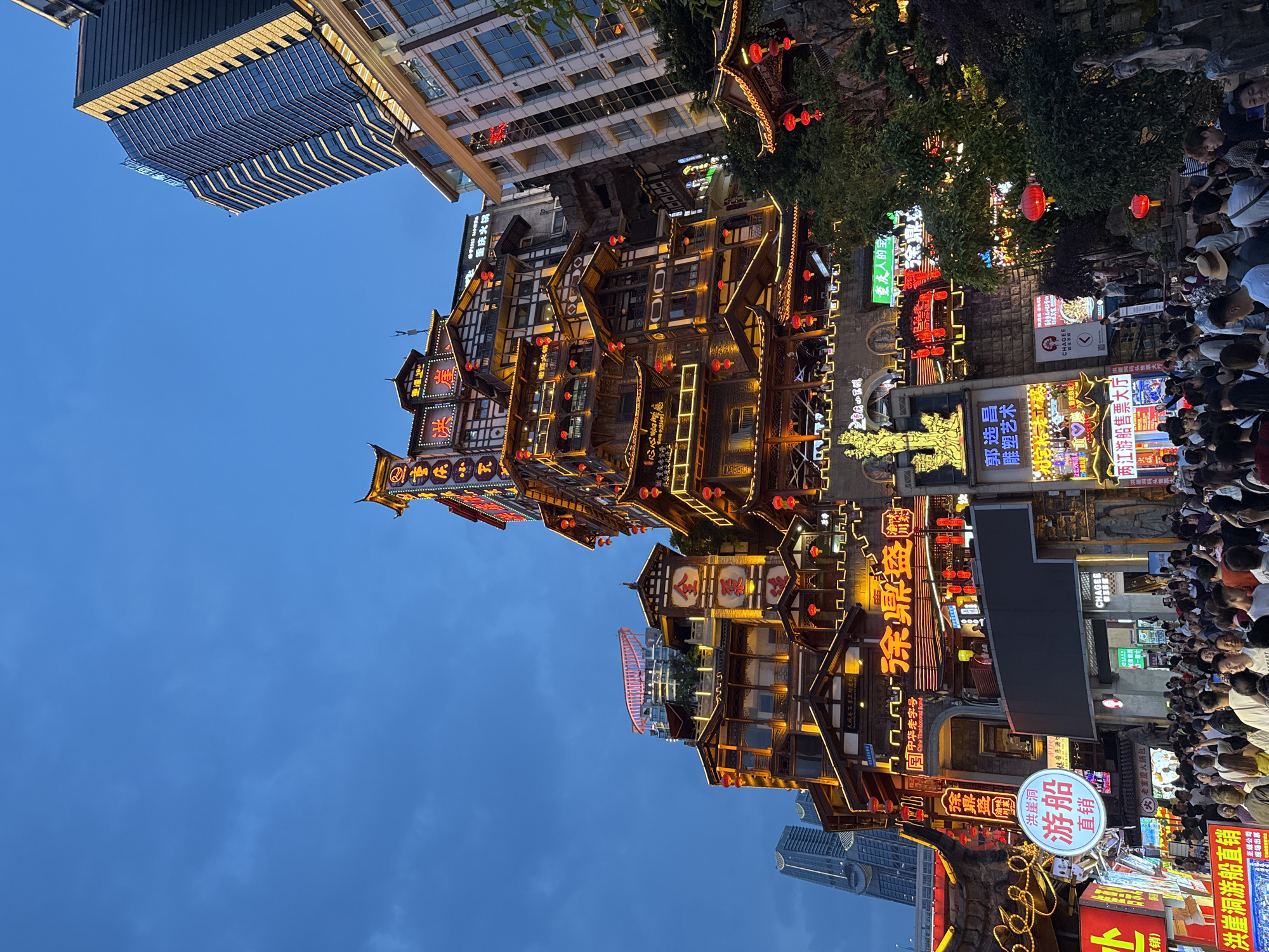 The image shows Hongyadong, a popular tourist attraction in the Yuzhong District of Chongqing, China. The architecture features traditional Chinese elements with multi-tiered wooden structures adorned with red lanterns and bright neon signs in Chinese characters. The buildings are lit up, suggesting it is dusk or night. Crowds of people are visible on the street level, indicating the location's popularity. Modern skyscrapers are visible in the background, contrasting with the traditional buildings.