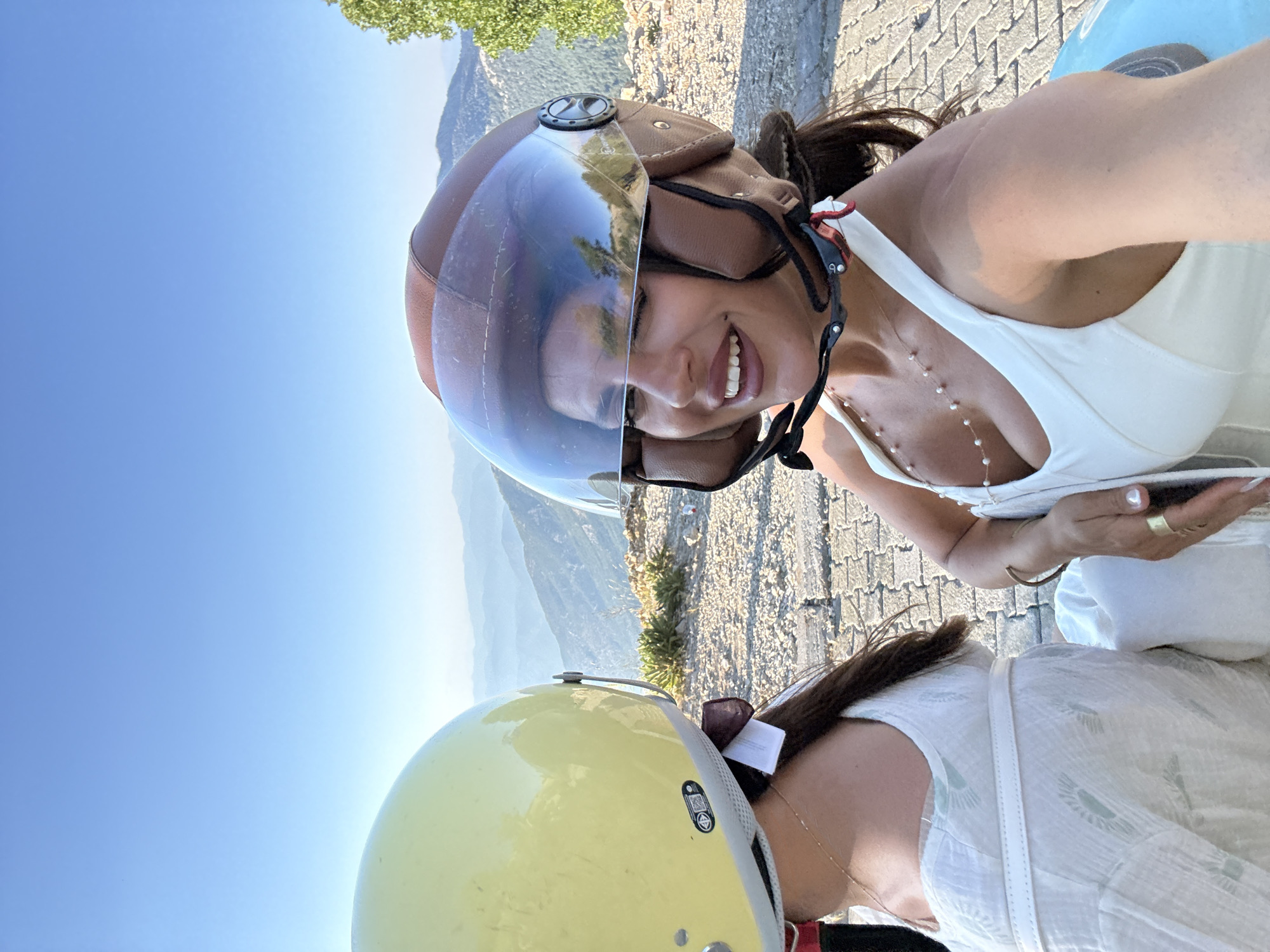 Two individuals wearing helmets are seated side-by-side, likely on a vehicle, with a scenic mountainous landscape in the background. The person on the right, in the foreground, is a woman wearing a brown helmet with a clear visor, a white top with a low neckline, and a pearl necklace. She is smiling and winking at the camera, suggesting a selfie or a posed photograph. Her right arm is visible, adorned with a gold ring. The person on the left is wearing a yellow helmet and a white dress with subtle patterned details. Their back is partially visible, and they appear to be looking forward. The setting is outdoors, on a paved surface, with a clear blue sky above. The mountainous terrain behind them is characterized by layers of hills receding into the distance, suggesting a location with elevated views, such as Ölüdeniz or Kayaköy, near Fethiye, Turkey.