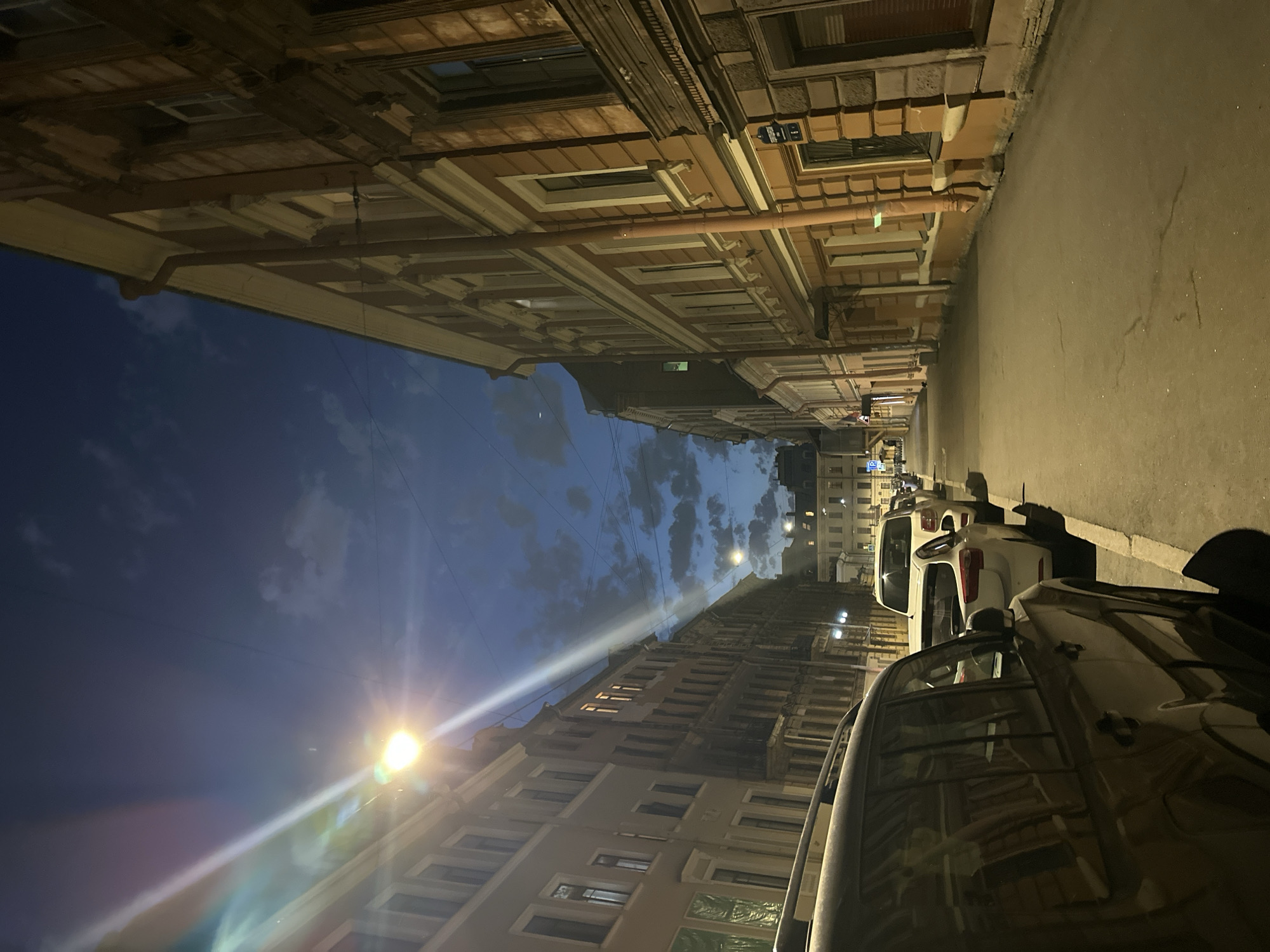 The image shows a narrow street in Saint Petersburg, Russia, at night. The street is lined with buildings of varying heights and architectural styles. A row of parked cars occupies the right side of the street. The sky is a mix of dark blue and clouds. A street lamp shines brightly, creating a lens flare. The angle of the image is slightly tilted.