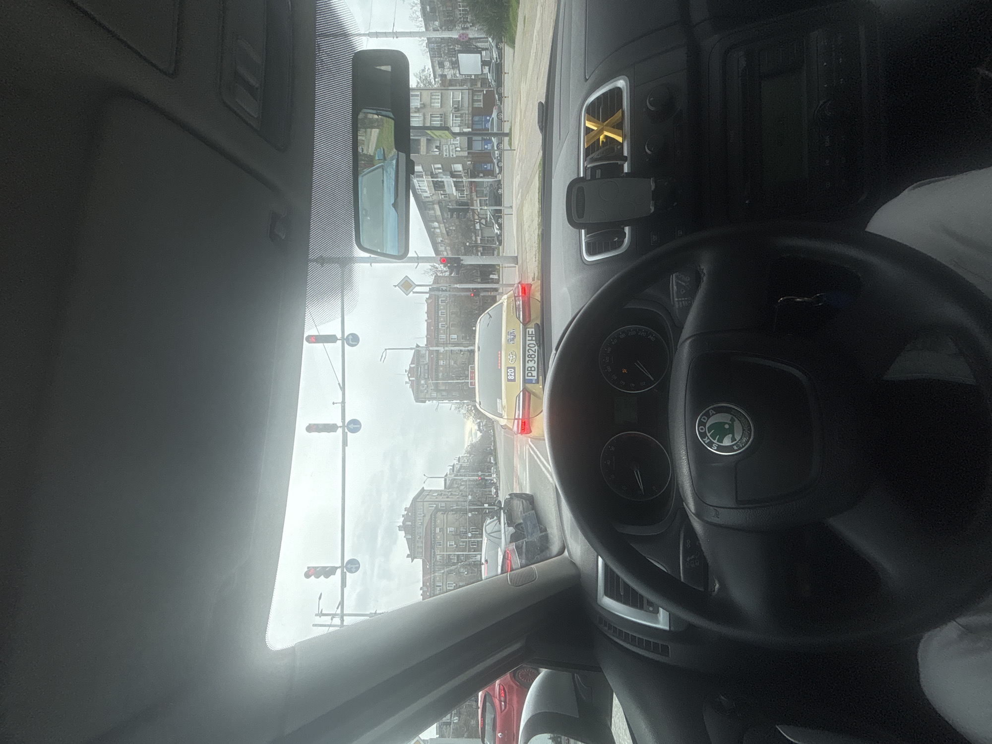 The image is taken from inside a car, likely the driver's seat, in Plovdiv, Bulgaria. The view through the windshield shows a street with buildings on both sides. The traffic light is red, and there are several cars ahead, including a yellow taxi with the license plate "PB 3820 HE." The steering wheel with a Skoda logo is visible in the foreground. There is a phone mount on the dashboard. The sky is overcast.