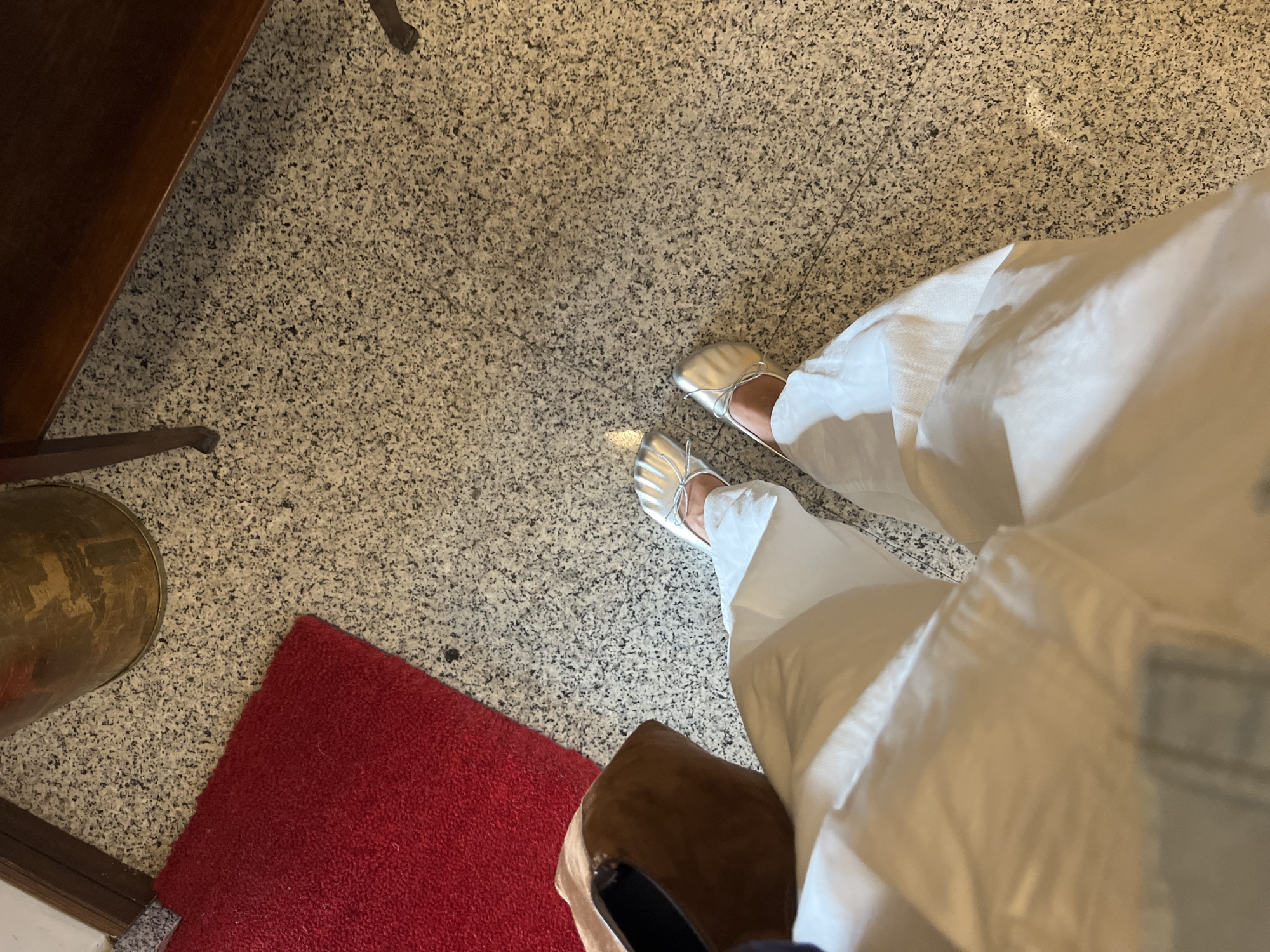 An overhead shot captures the lower body of a person standing indoors in Milan, Italy. The individual is wearing white, wide-leg trousers and silver ballet flats adorned with small bows on the toes. The floor is a light-colored granite-patterned tile with black and grey speckles, and a visible seam runs through it.
In the upper left quadrant, a dark brown wooden furniture item, possibly a table or chair leg, is partially visible. Towards the lower left, a corner of a bright red, textured rug is laid on the floor. Adjacent to the rug, a curved, brass-colored object, resembling a decorative pot or container, is also partially visible. Another dark brown object, possibly a shoe or bag, is glimpsed in the lower right.
The lighting is bright and even, suggesting an indoor setting. The image focuses on the interplay of textures and colors, particularly highlighting the silver footwear against the patterned floor and white clothing. No text is visible in the image.