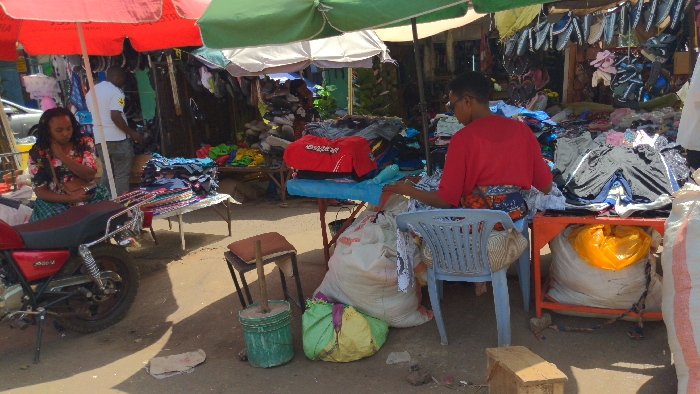 This image captures an outdoor market scene in Moshi, Tanzania, under bright daylight. In the left foreground, a woman with dark hair, dressed in a floral top and a plaid skirt, is seated sideways on a red motorcycle, looking down. Behind her, a man in a white t-shirt stands partially visible near a display. To the immediate right of the motorcycle, a table holds multiple stacks of folded garments. A red market umbrella provides shade over this section.
In the center, a small red table displays a red garment and other clothing items. On the ground nearby, a green bucket contains a wooden implement, and a small cushioned stool is present. Several large sacks, including white and green ones, are situated on the dirt ground.
On the right, a woman wearing a red shirt and a patterned skirt is seated on a light-blue plastic chair, facing away from the viewer. She is actively sorting through clothing items arranged on an orange table in front of her. A large white sack with a yellow liner is positioned beneath this table. A green market umbrella provides overhead shade on the right.
The background features numerous market stalls with various clothing items piled on tables and hanging from makeshift structures. The ground throughout the scene is composed of packed dirt or gravel.